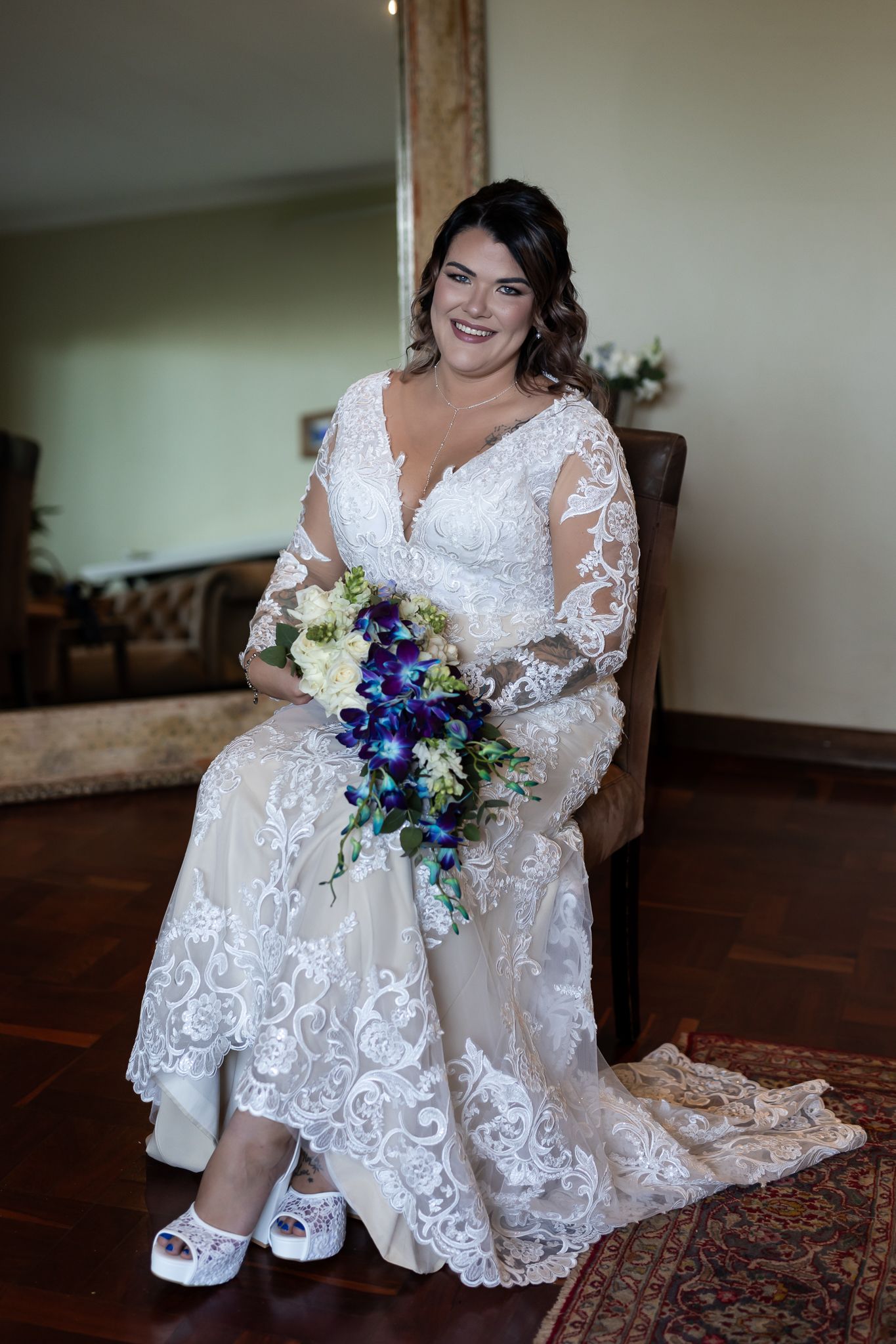 Bride sits elegantly in lace wedding dress holding purple and white bouquet.