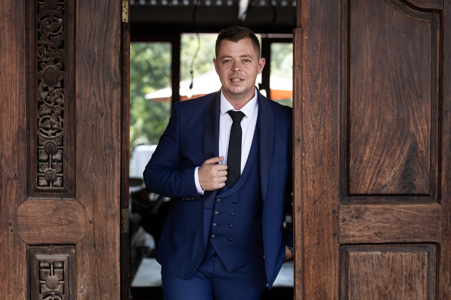 Man in blue suit standing in ornate wooden doorway while adjusting his tie.