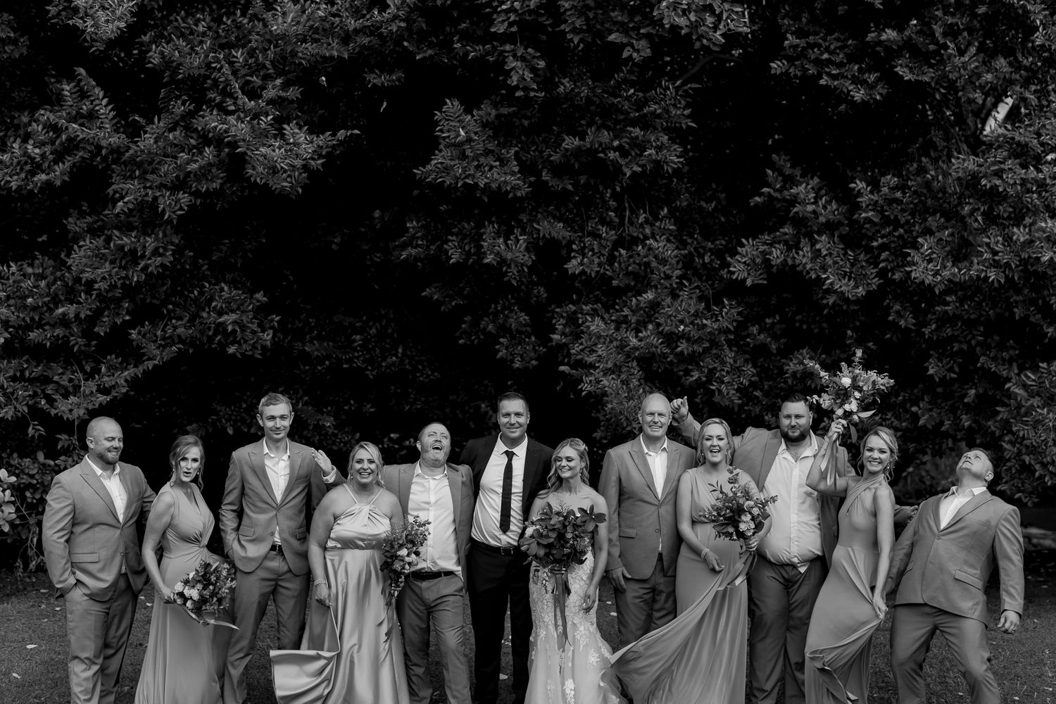 Black and white portrait of wedding party lined up against dark foliage backdrop.
