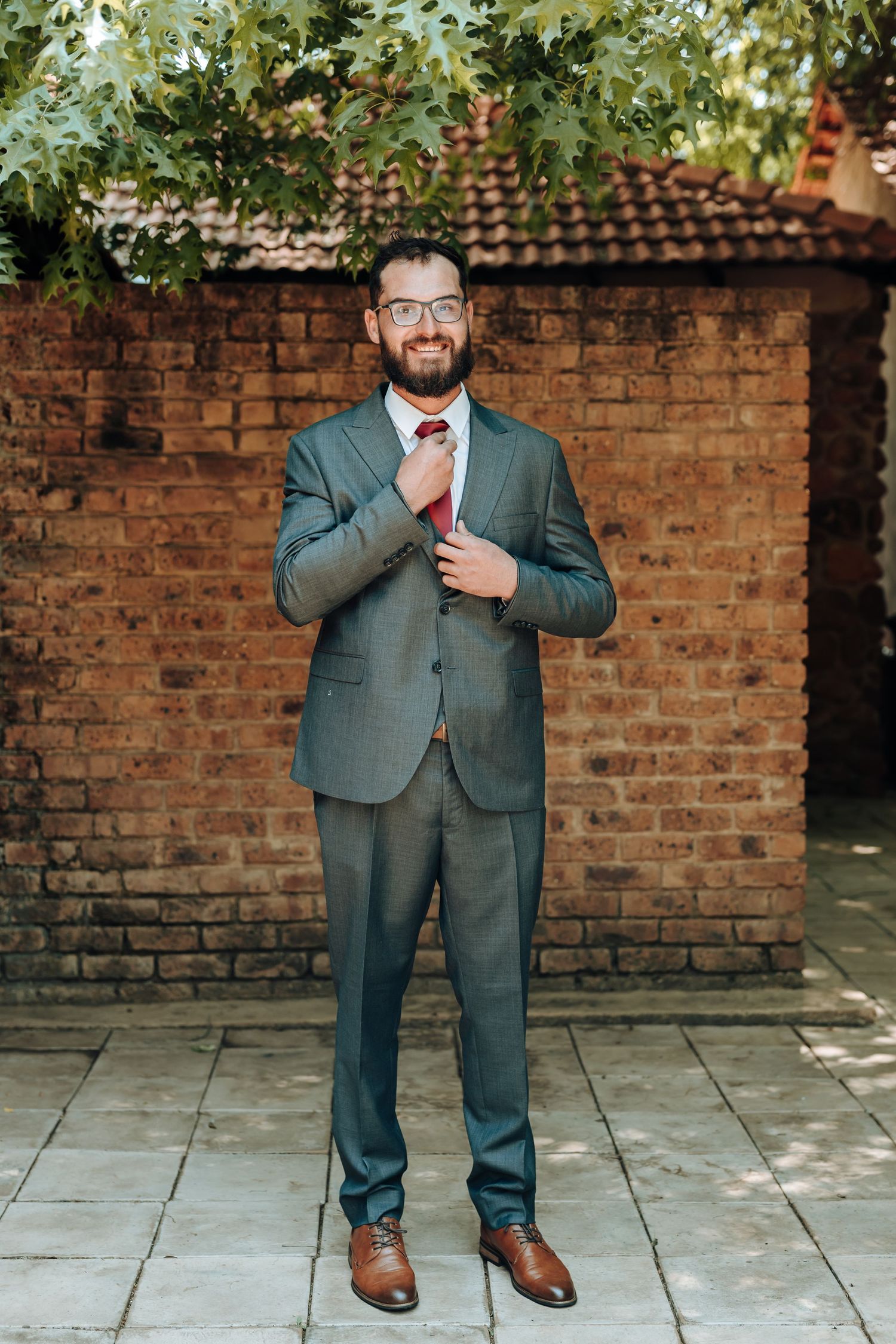 Professional standing portrait in grey suit against outdoor brick wall setting.