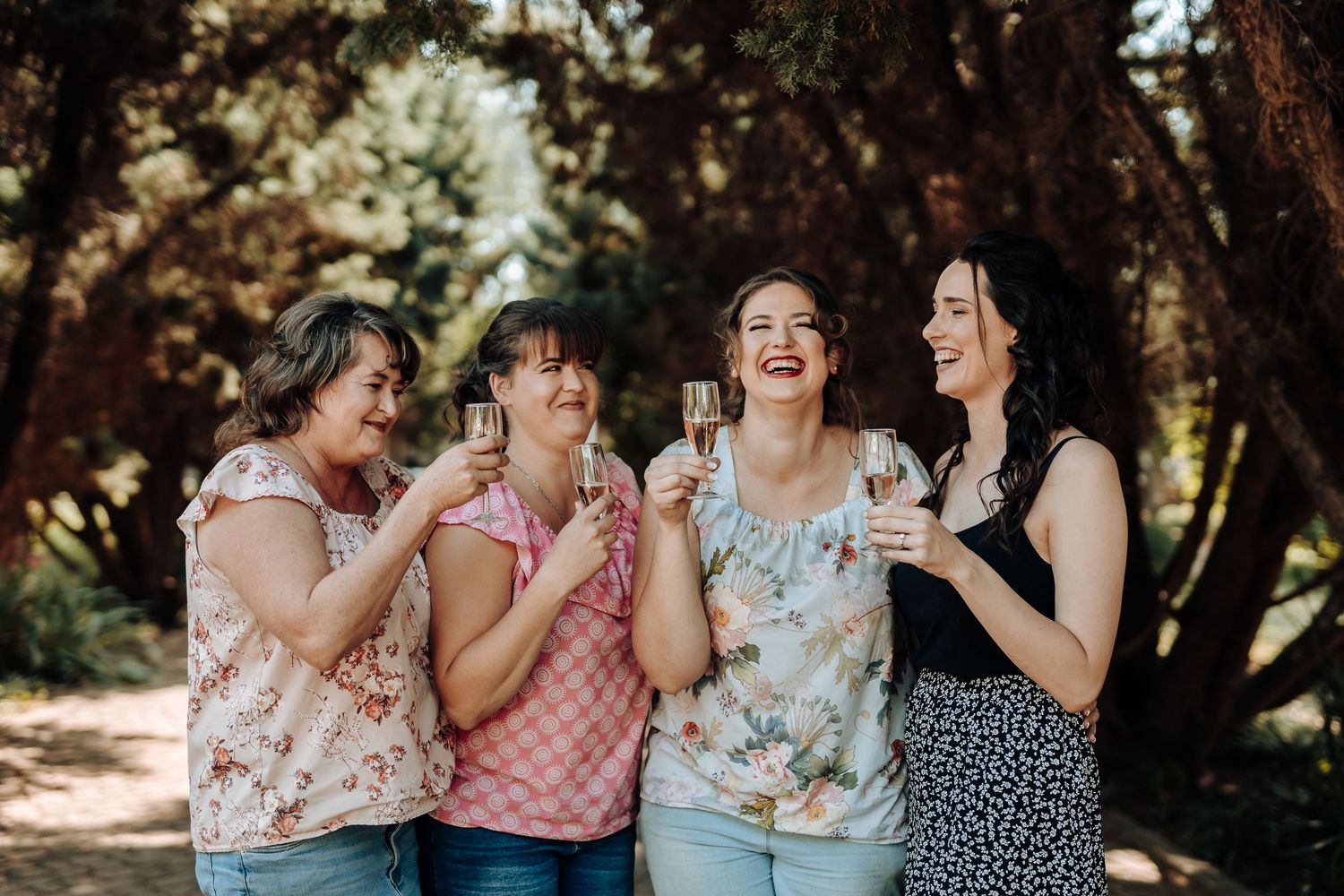 A group of friends laughing and holding champagne glasses while enjoying a celebration in a wooded outdoor setting.