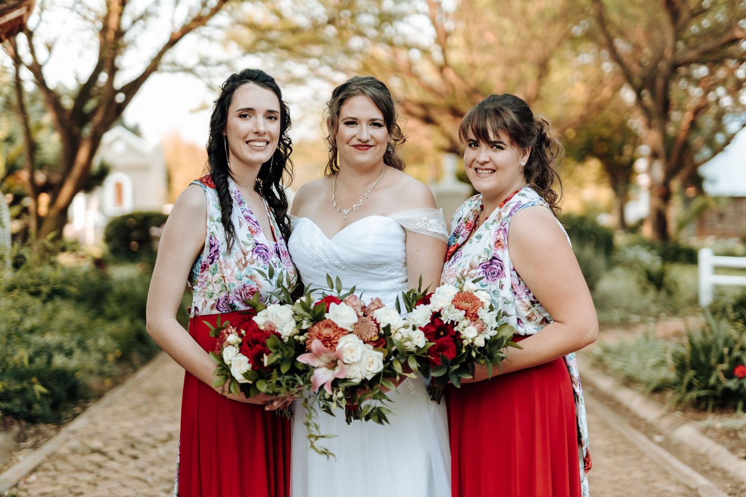 Three women in red and floral dresses pose with beautiful bouquets at an outdoor wedding.