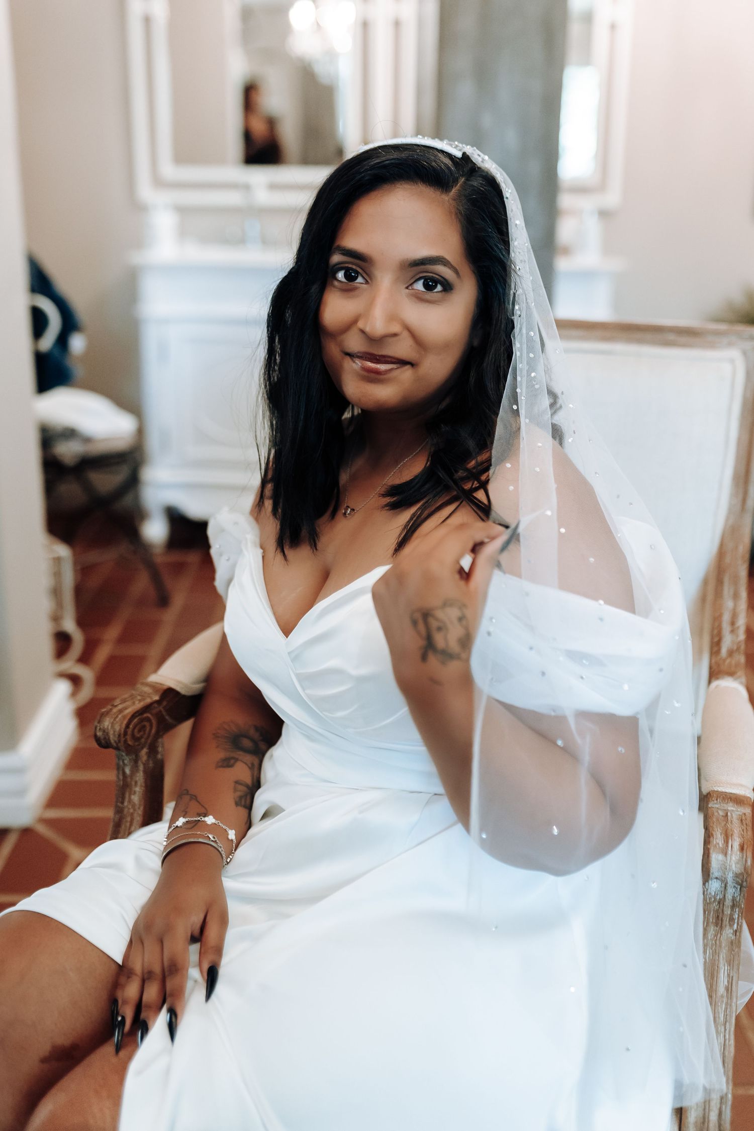 Bride in white off-shoulder wedding dress with long dark hair and veil poses elegantly in an indoor setting.