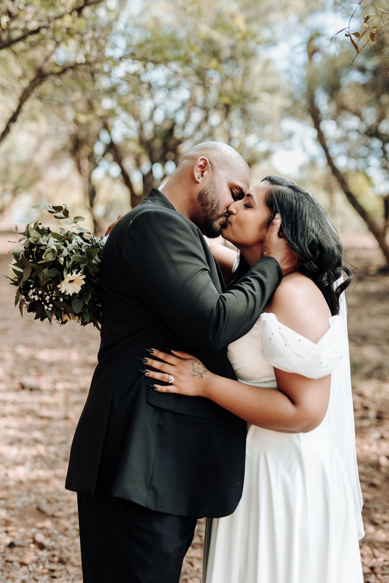 Romantic wedding couple shares a kiss while holding floral bouquet in wooded setting.