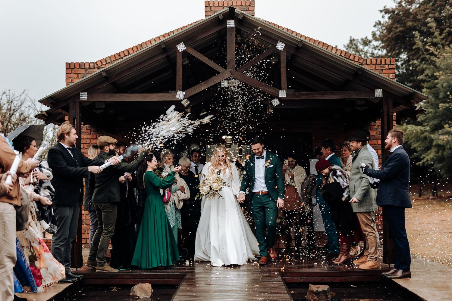 Joyful wedding party celebrates with confetti toss outside wooden pavilion on overcast day.