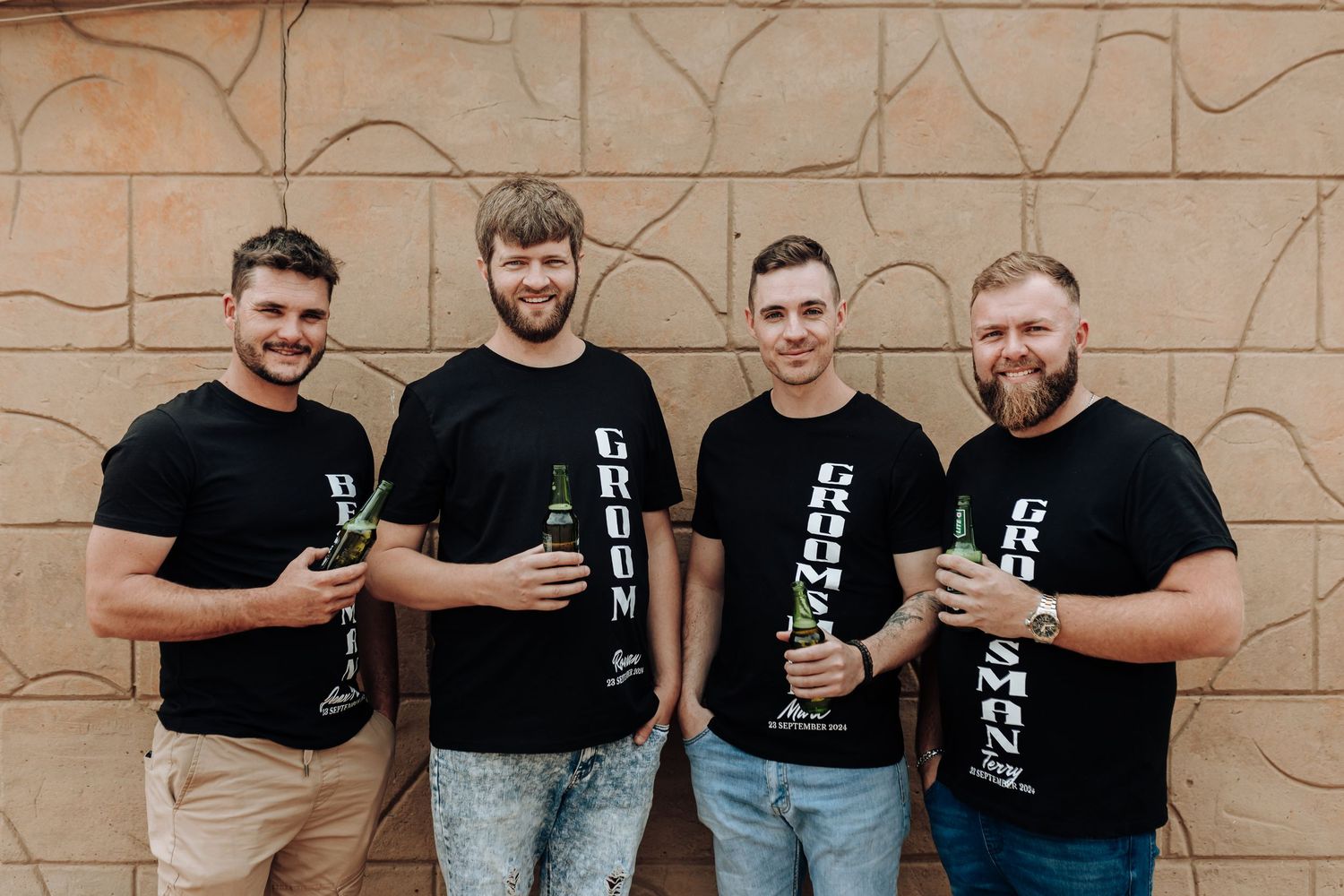 Group of four people in matching black t-shirts smile while posing against wall.