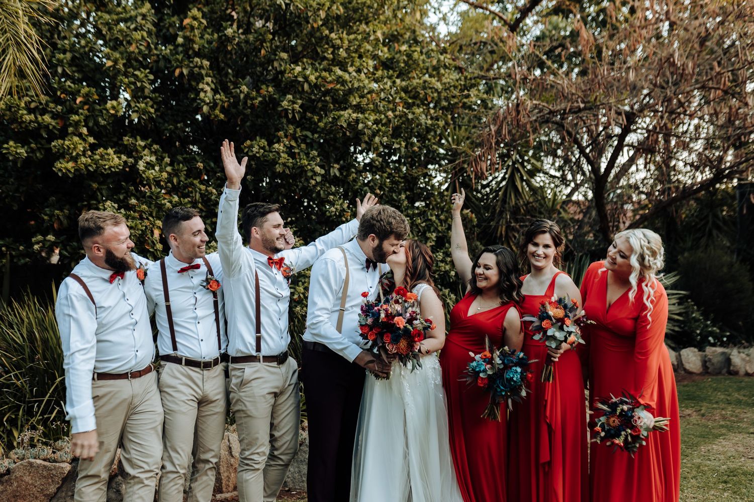 Joyful wedding party celebrating with raised arms outdoors in garden setting.