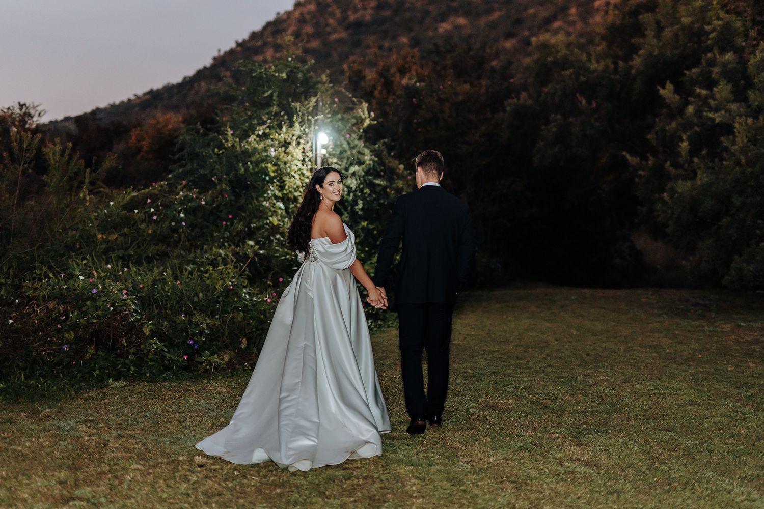 A romantic wedding couple stands in a garden at dusk with sparkling lights creating a magical atmosphere.