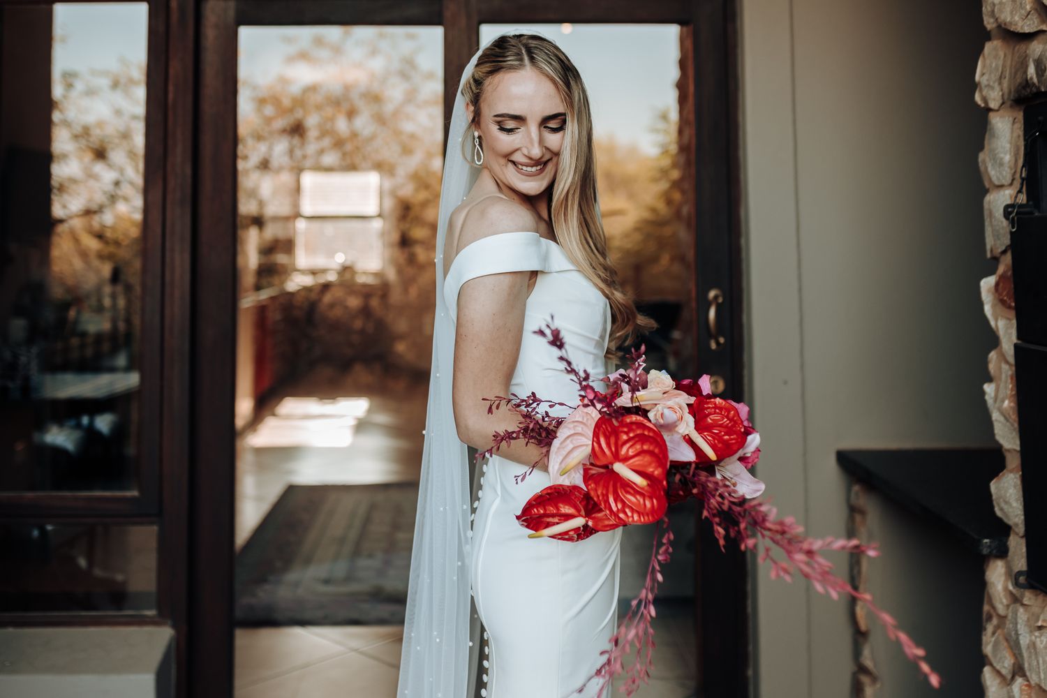 Bride in sleek white gown smiles while holding dramatic red floral arrangement.