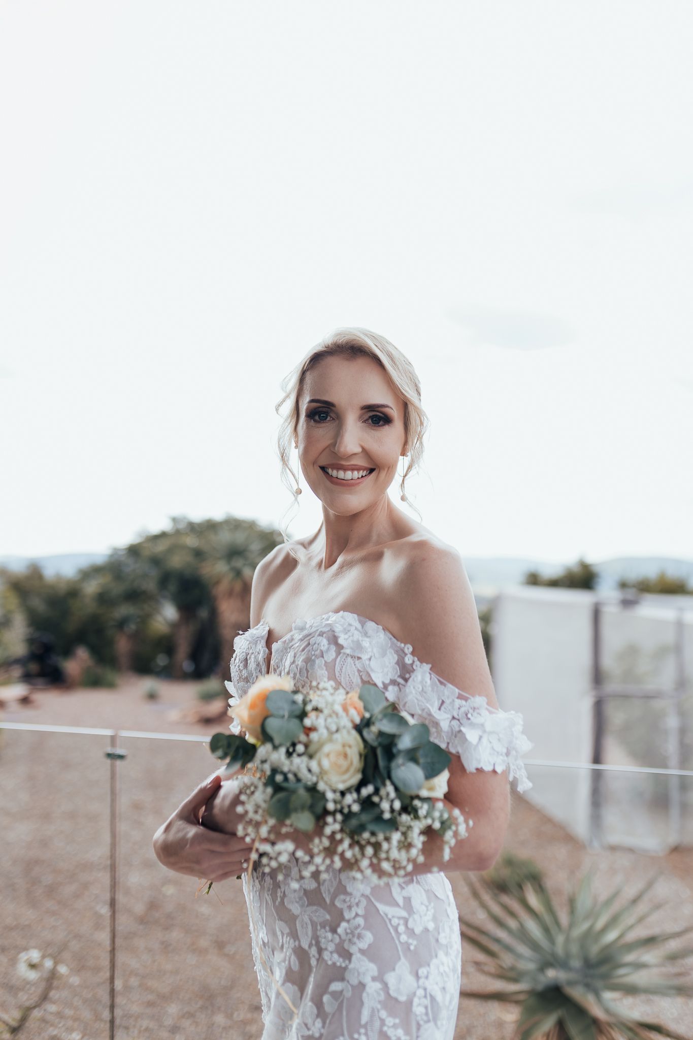 A bride smiles while holding her bouquet on an outdoor deck with scenic mountain backdrop.