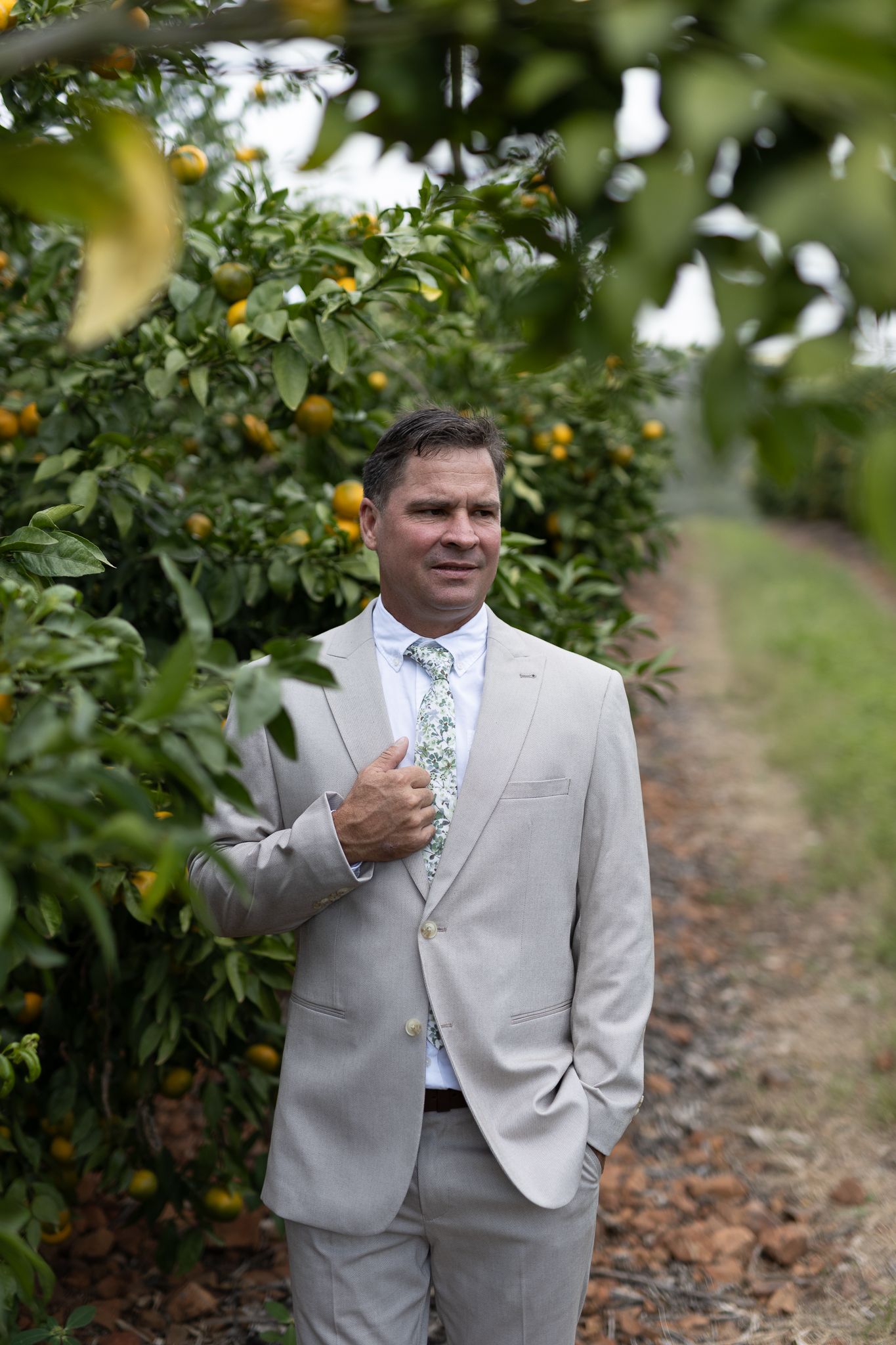 Wedding attendee in light gray suit poses among fruit trees in an orchard.