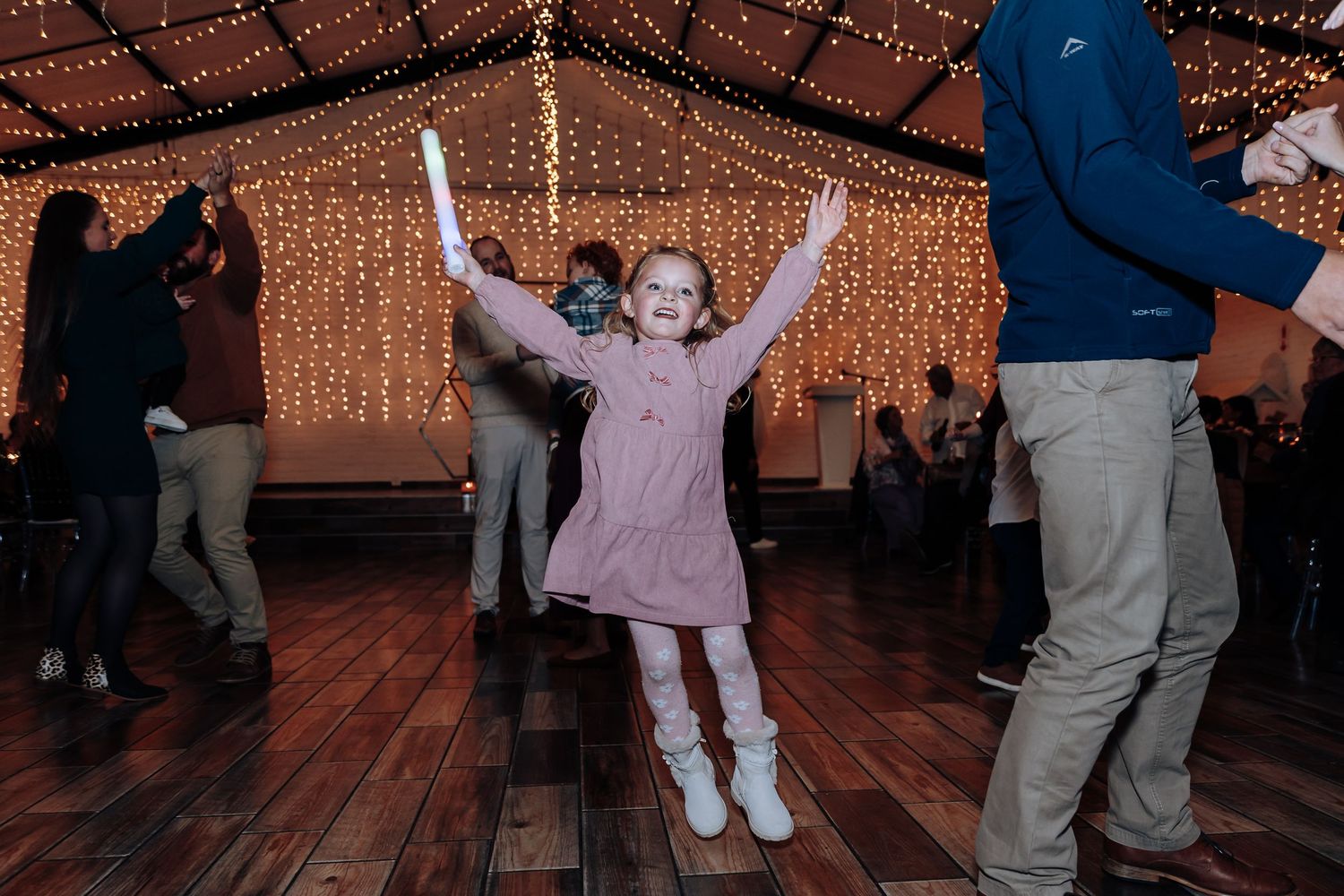 Young girl dancing joyfully at a wedding reception under fairy lights on a wooden dance floor.