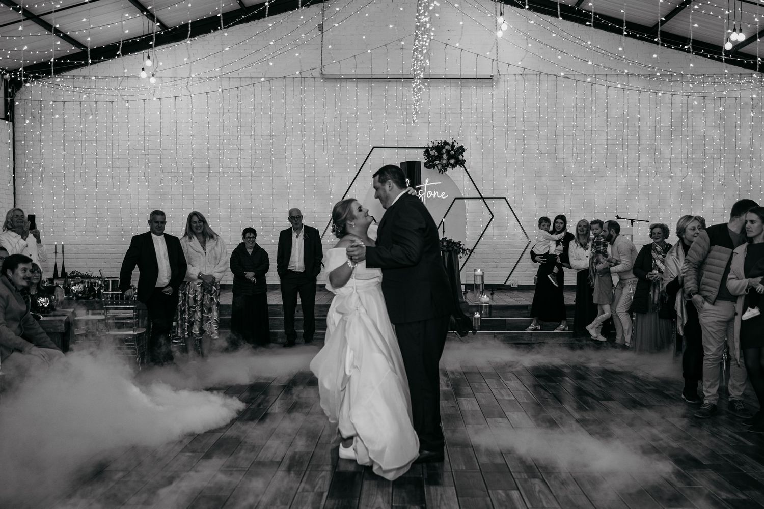 Black and white photo of a couple sharing their first dance at a wedding reception with fog effects and fairy lights.