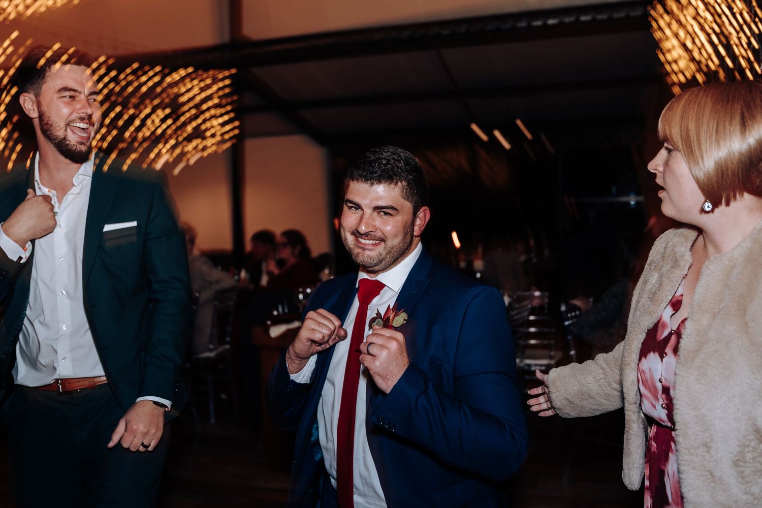Man in blue suit and red tie smiling at a wedding reception party with guests in background.