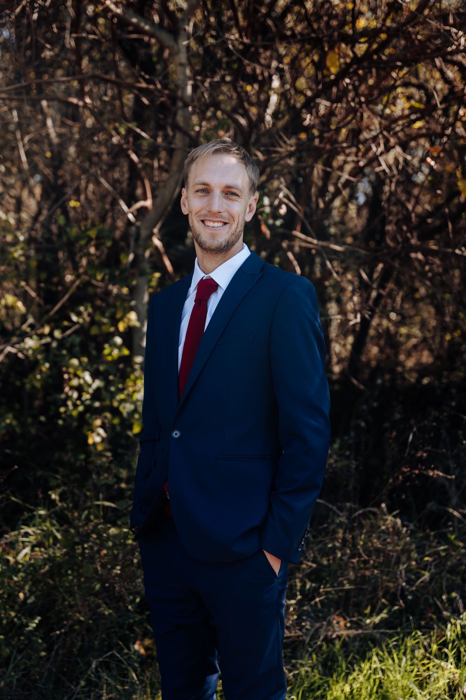 Man in navy blue suit and red tie smiling outdoors with autumn trees in background.