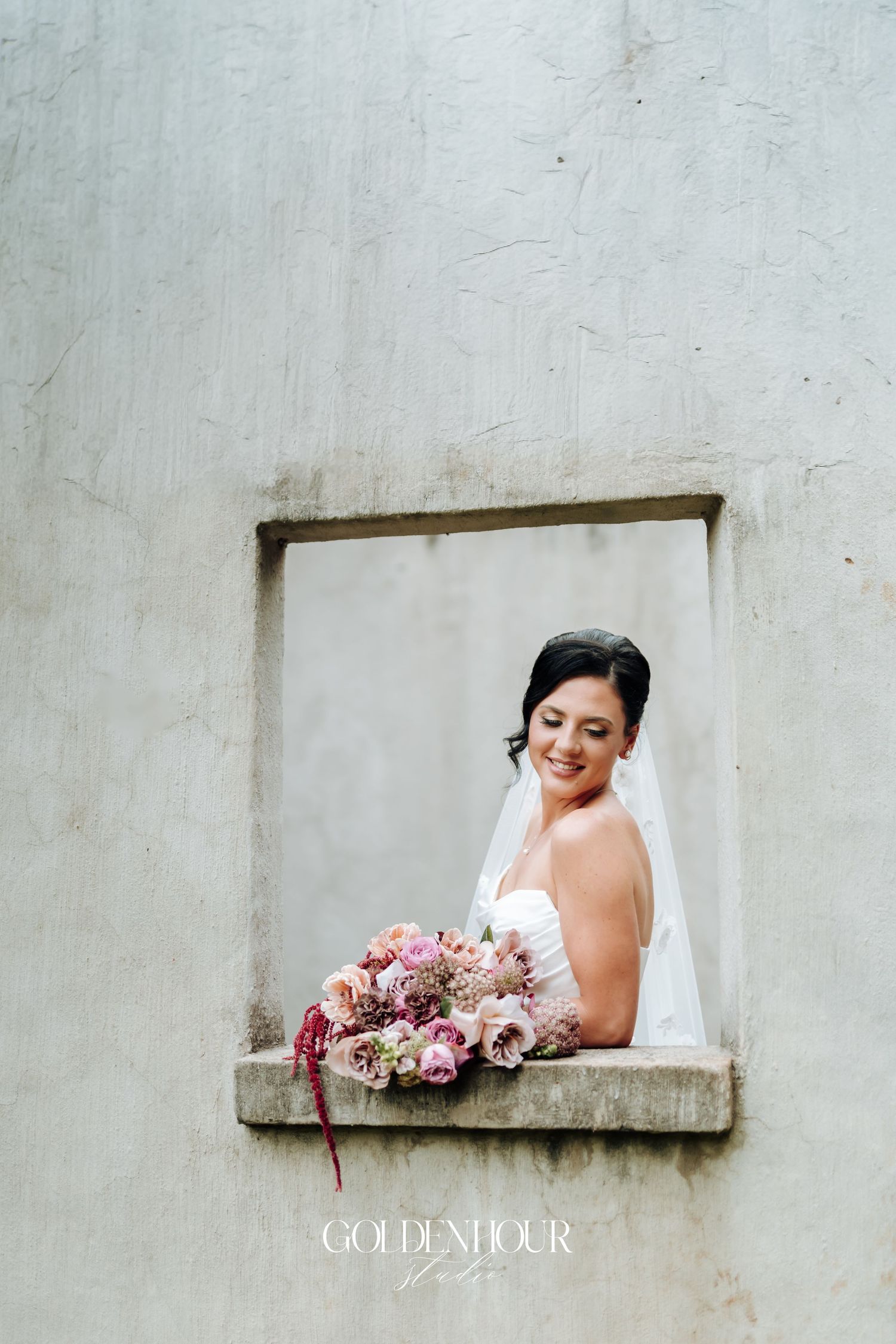 Radiant bride in white gown and veil holding a pink bouquet, posing in a rustic stone window alcove.