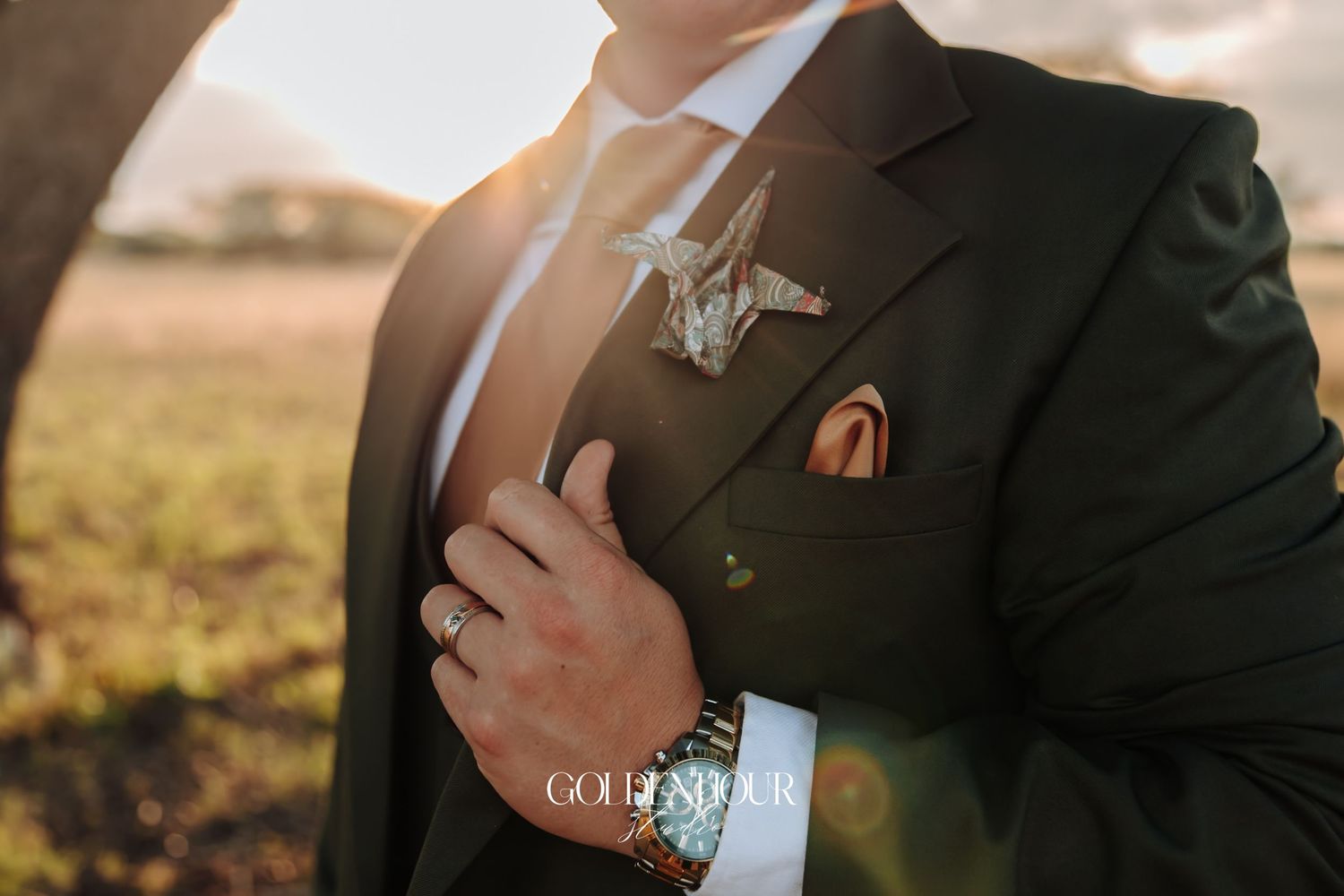 Close-up of groom adjusting dark suit with boutonniere in golden sunset light.