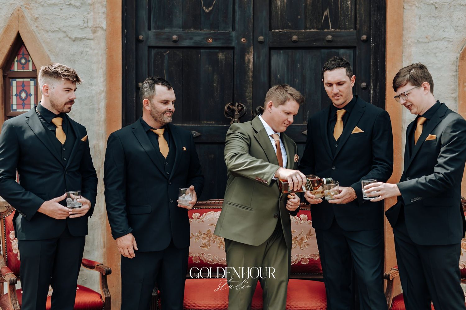 Groom and groomsmen in black suits with gold ties sharing a toast with whiskey glasses before the wedding.