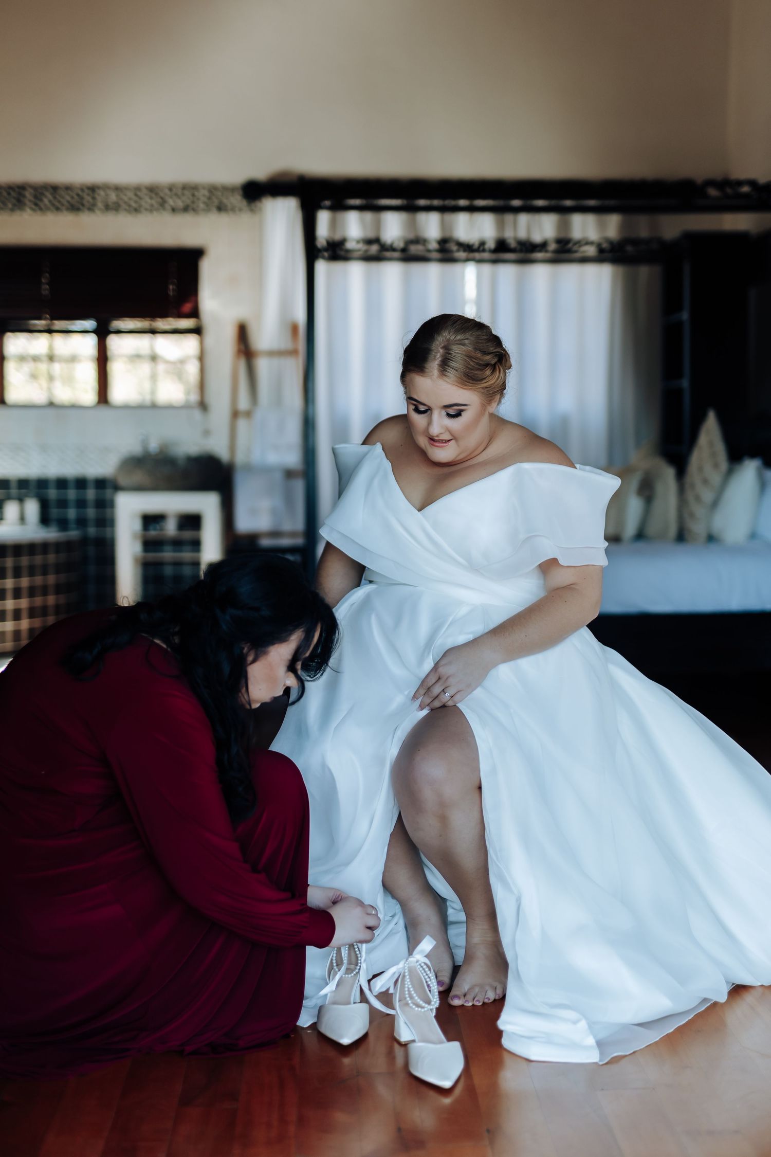 Bridesmaid in red dress helps bride in white off-shoulder gown put on white heels in elegant bedroom.
