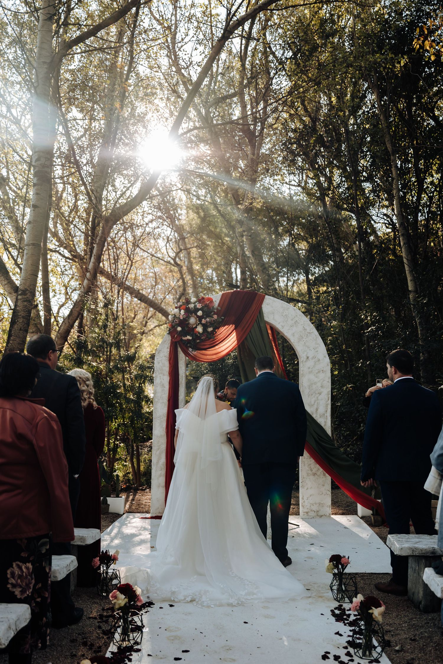 Bride and groom stand at floral arch decorated with burgundy draping in sunlit forest wedding ceremony.