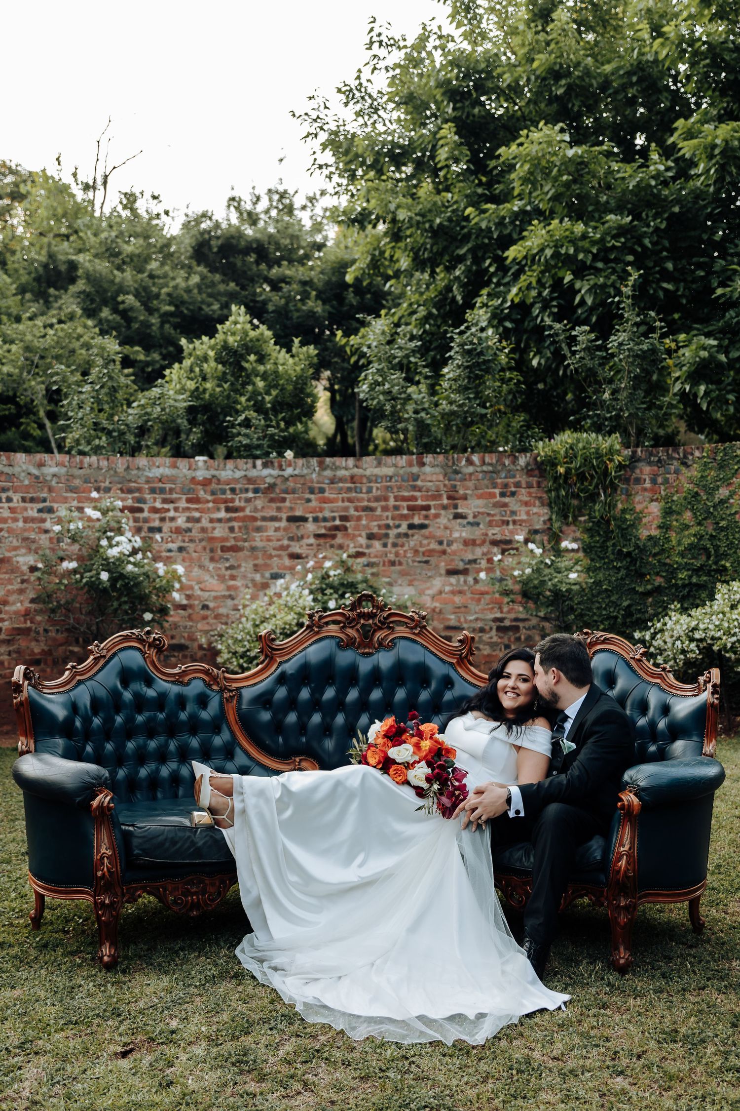 Wedding couple lounging on vintage blue tufted couch outdoors against brick wall.