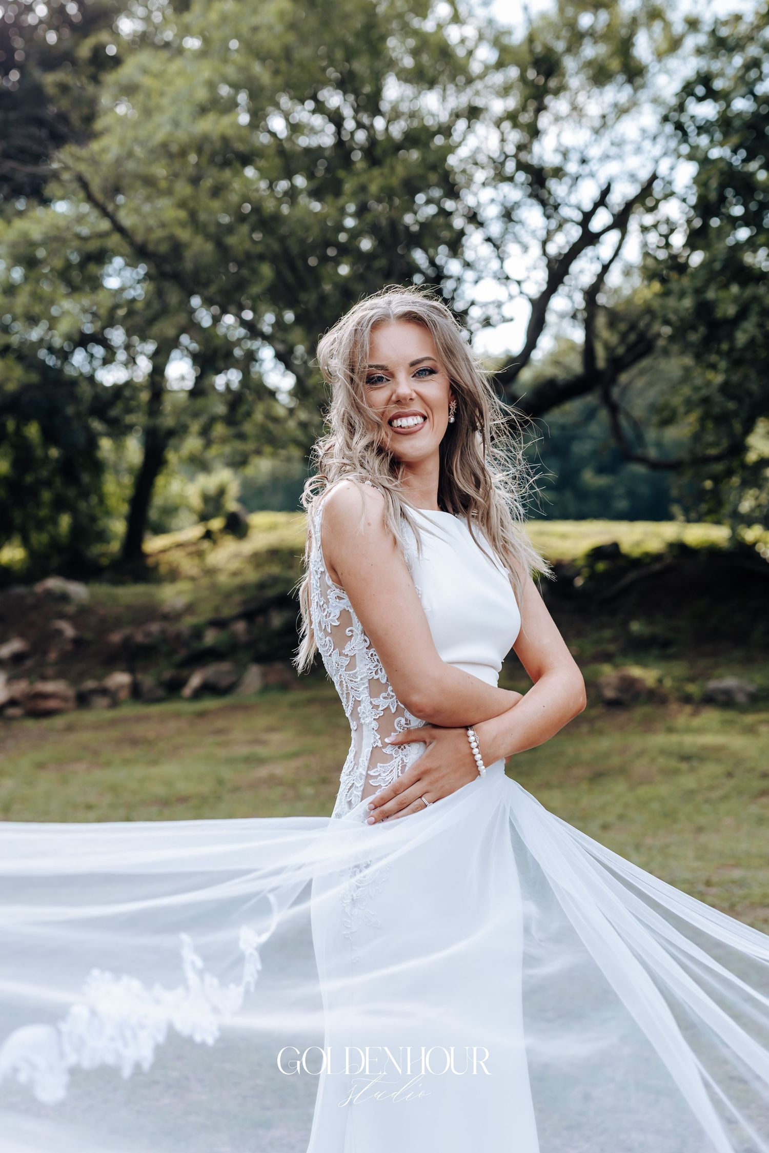 A flowing white wedding dress is showcased in a natural outdoor setting with trees and greenery.
