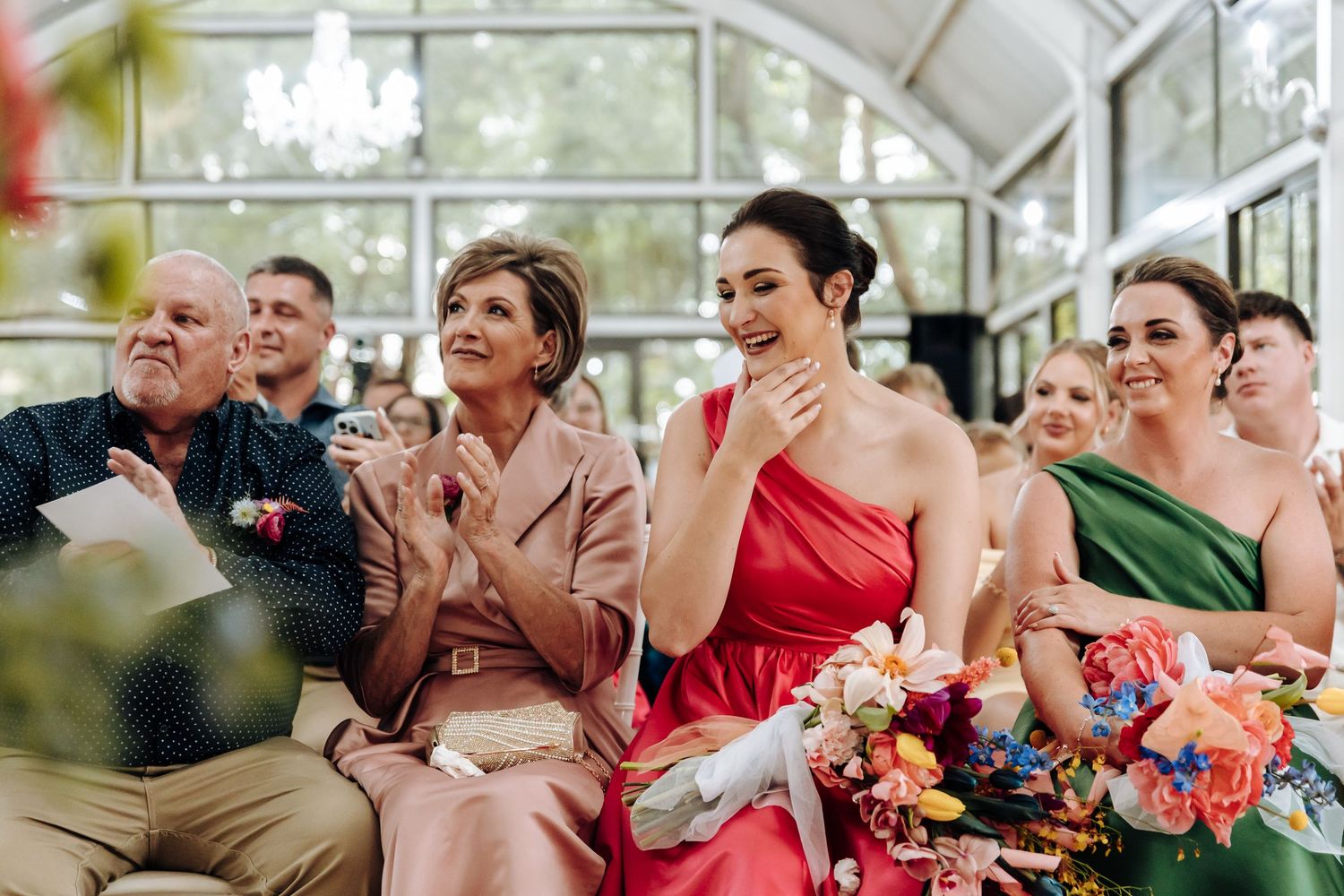 Wedding guests react emotionally during a ceremony in a bright sunlit conservatory with colorful floral decor.