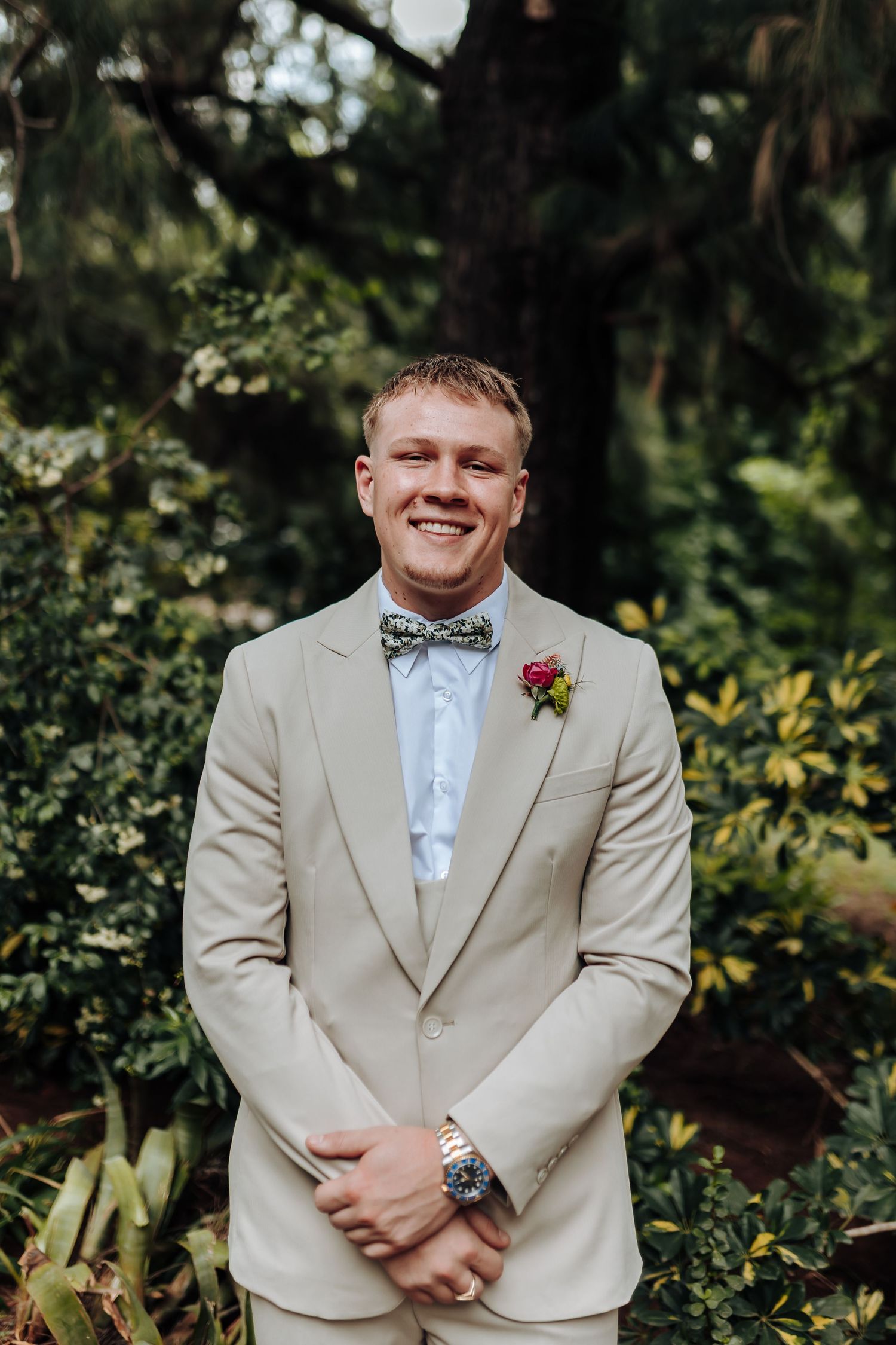 A person in a beige suit and bowtie smiles while standing in a lush garden setting.