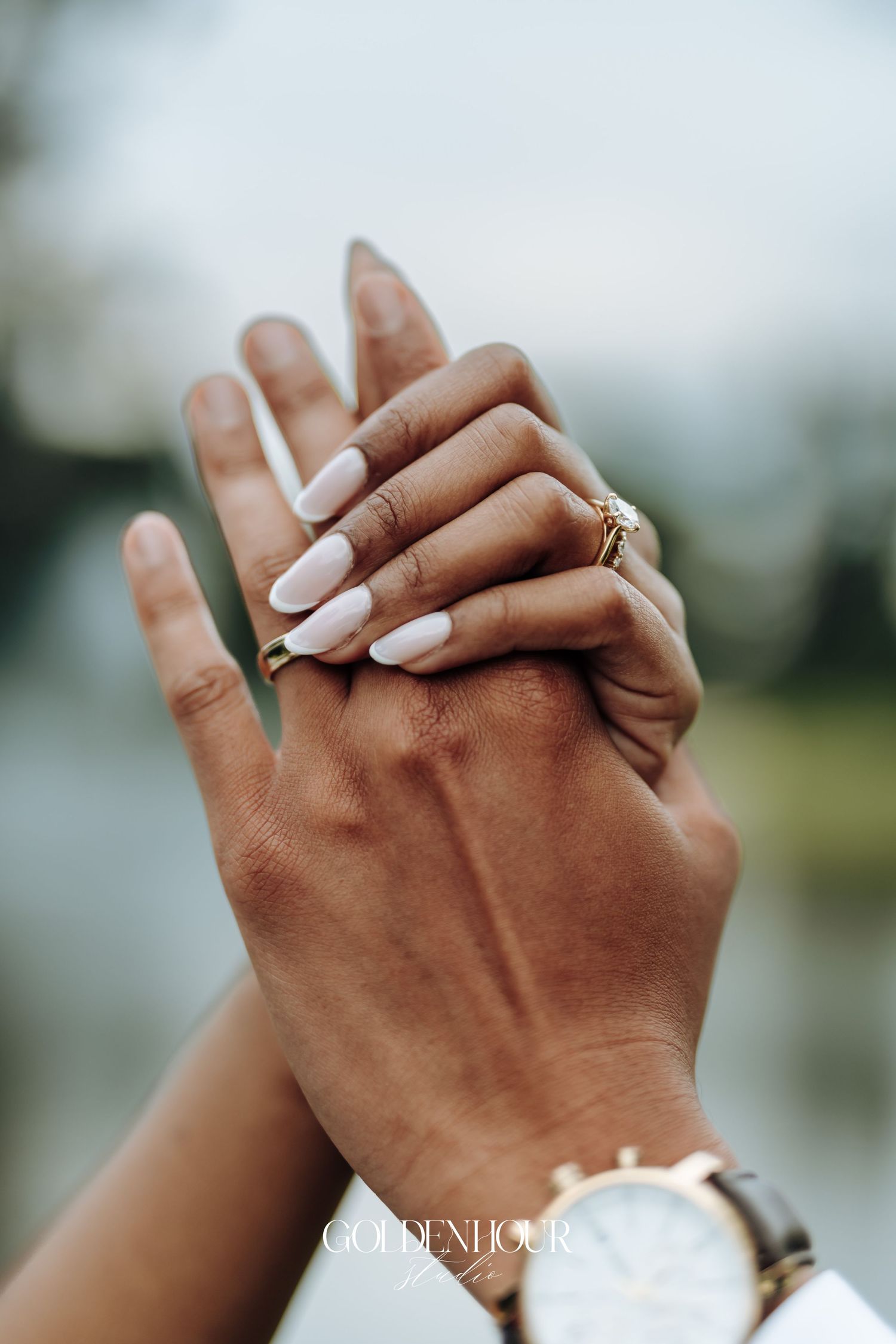 Close-up of clasped hands showing wedding rings against a blurred outdoor background.