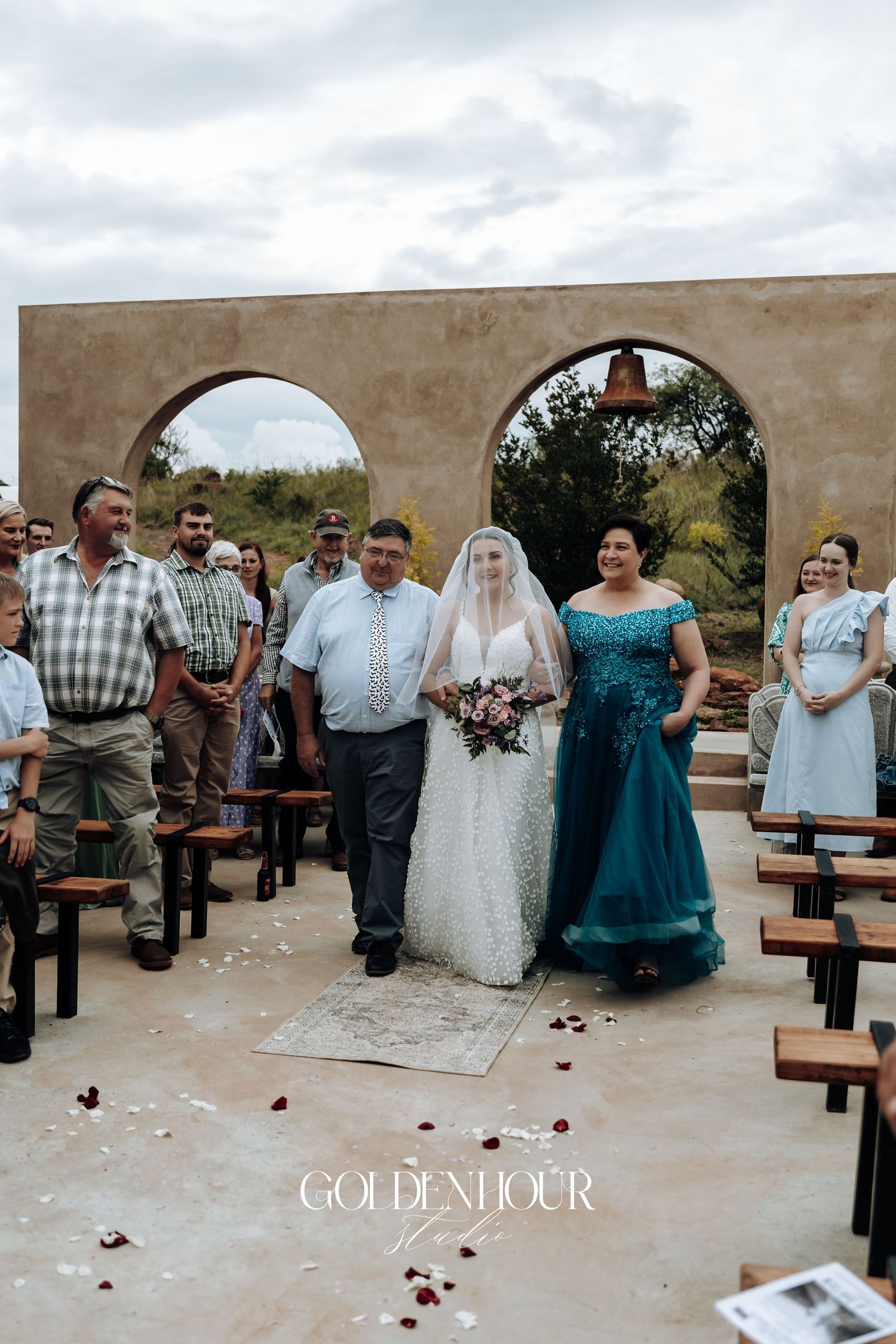 Outdoor wedding ceremony under stone arches with guests seated on wooden benches.
