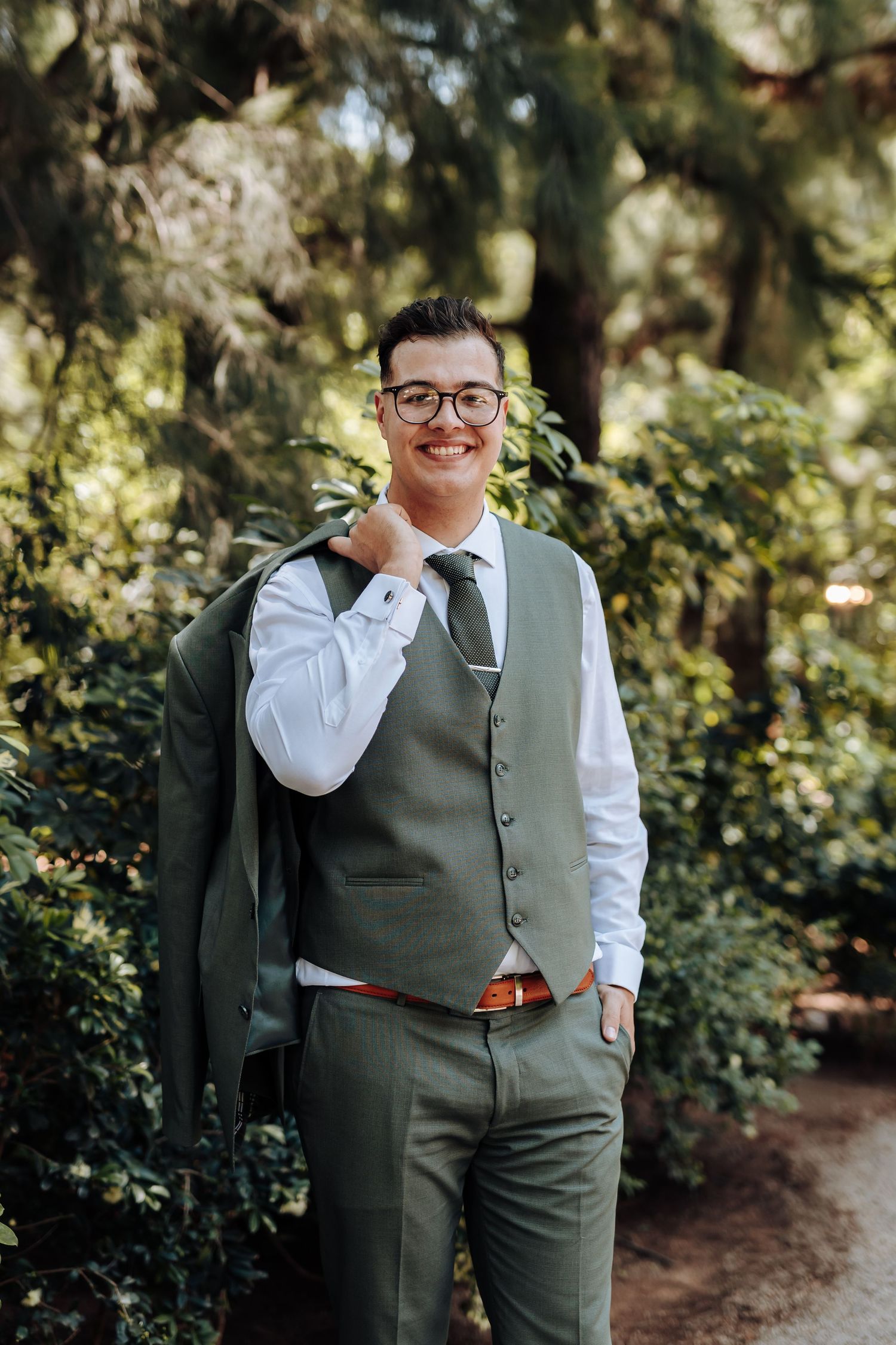 A person in a green vest and white dress shirt smiles while standing in a lush garden setting.