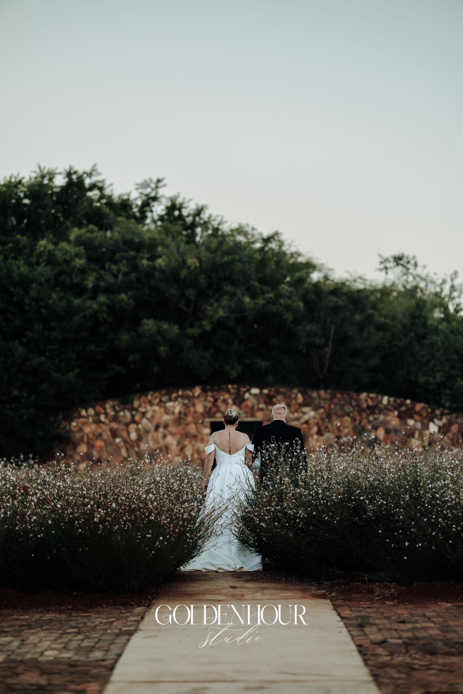 A bride in a white wedding dress sits on a wooden dock surrounded by wildflowers at sunset.