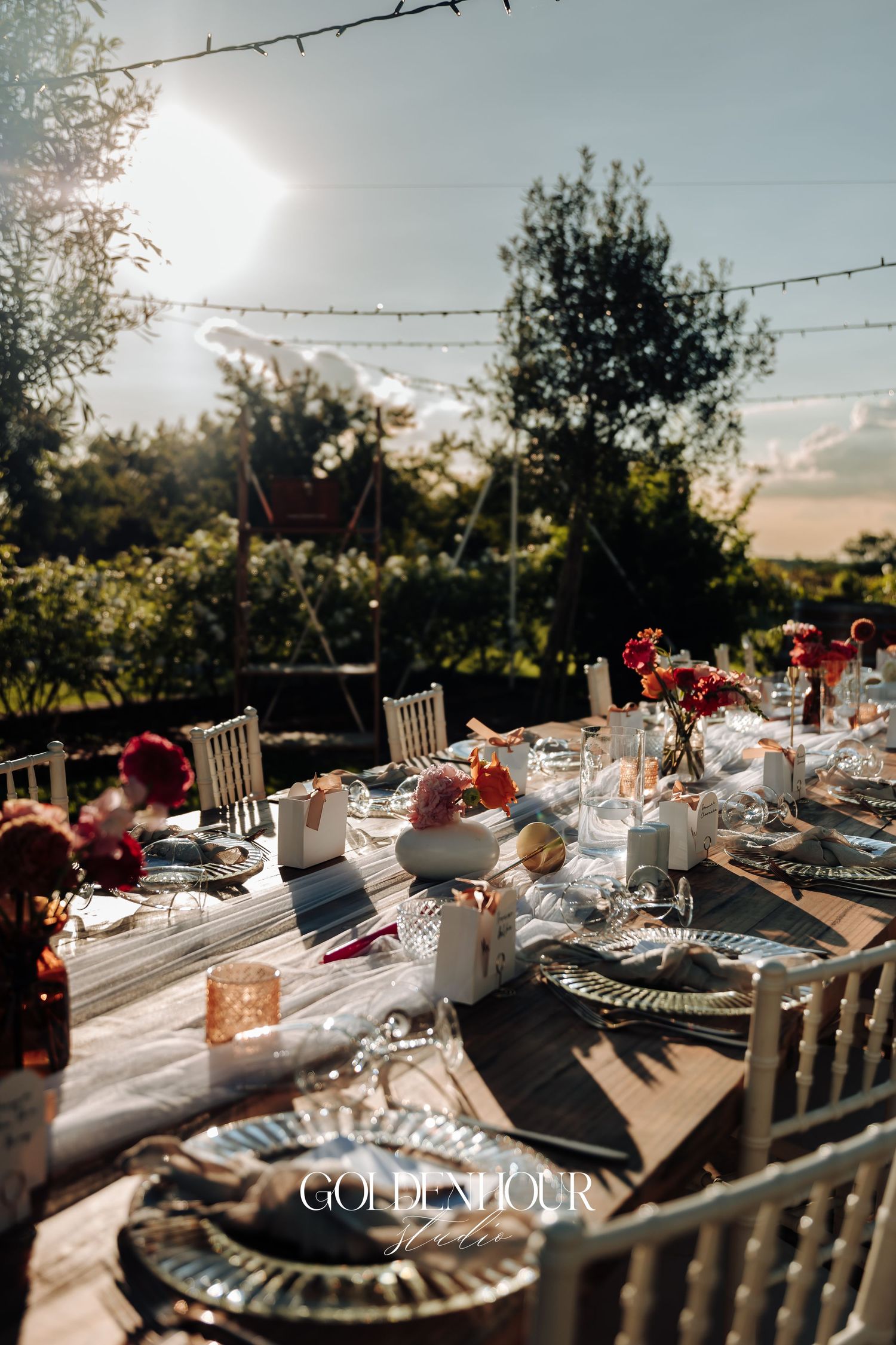 Elegant outdoor dining table settings with silver charger plates and red floral centerpieces at sunset.