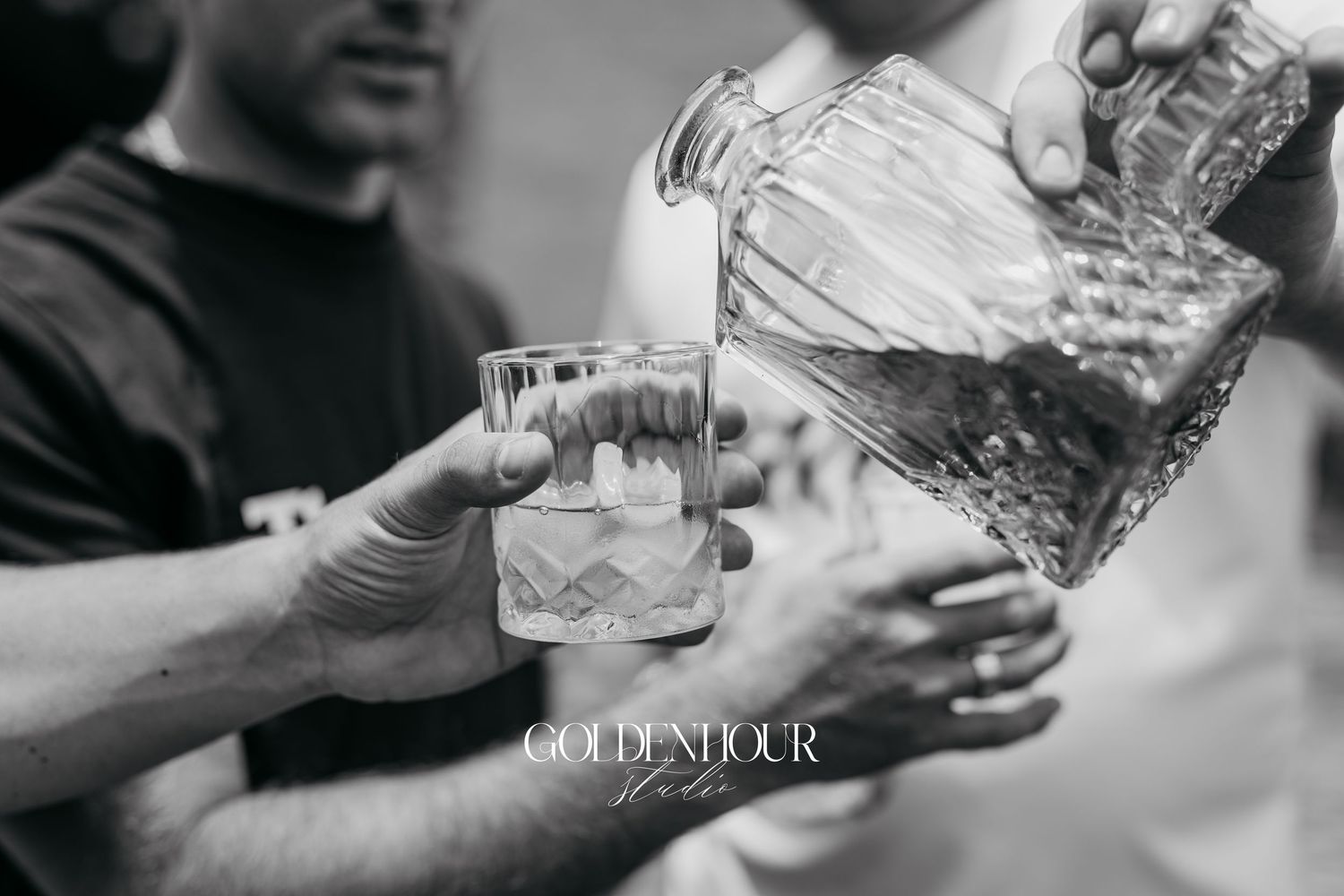 Hands pouring clear liquid from a glass bottle into a cocktail mixing glass in a dimly lit bar setting.