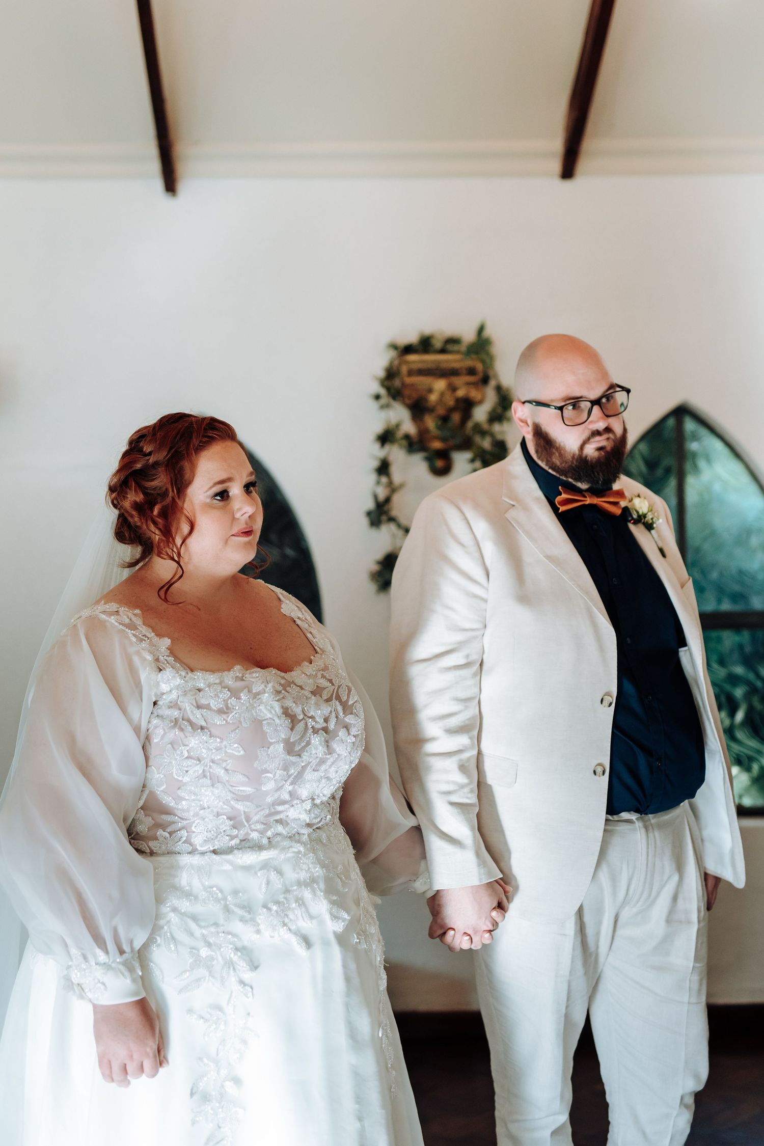 A couple in wedding attire holds hands while standing in a light-filled chapel with white walls and stained glass windows.