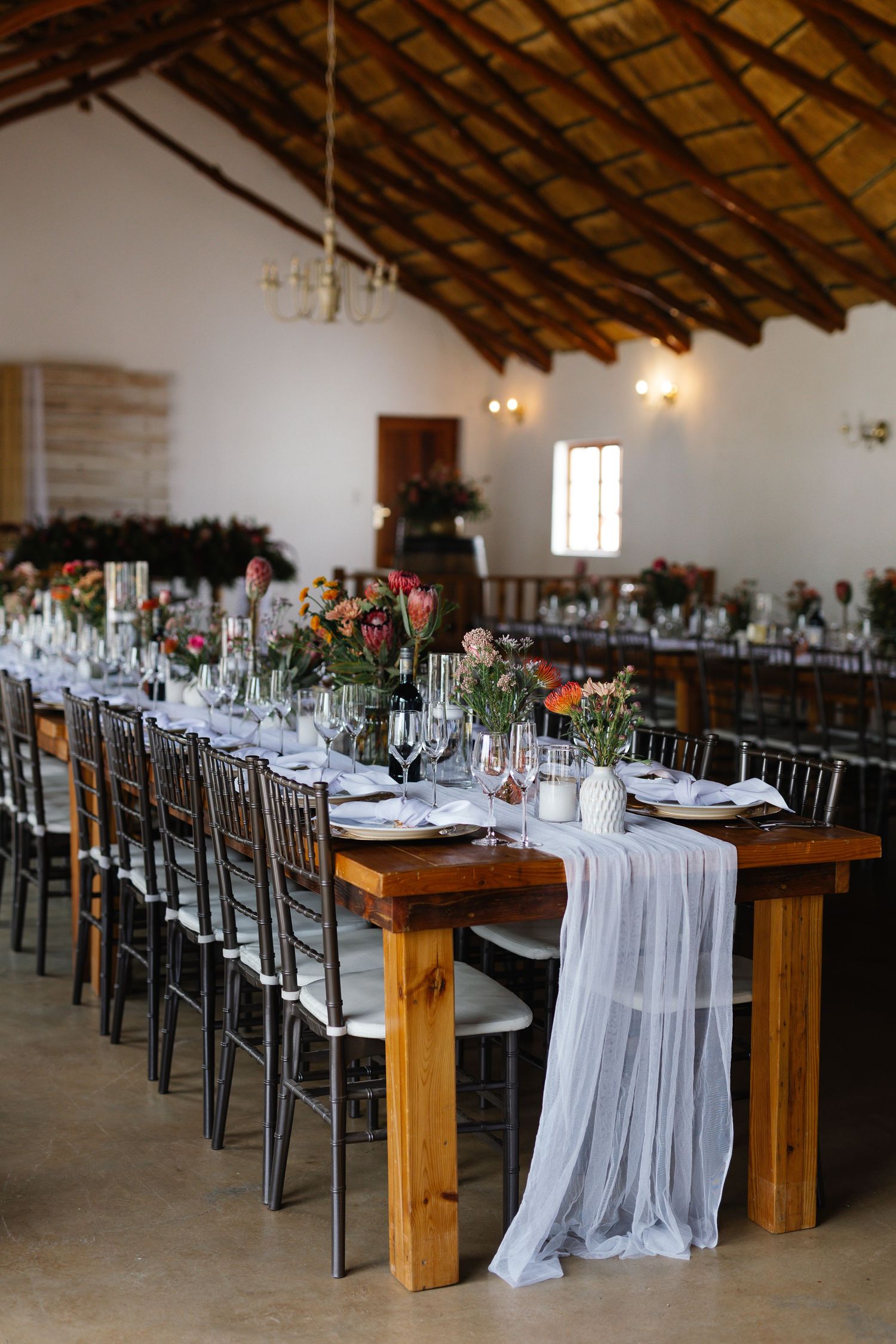 Long rustic wooden tables with white linens and metal chairs set up for an elegant wedding reception in a barn venue.