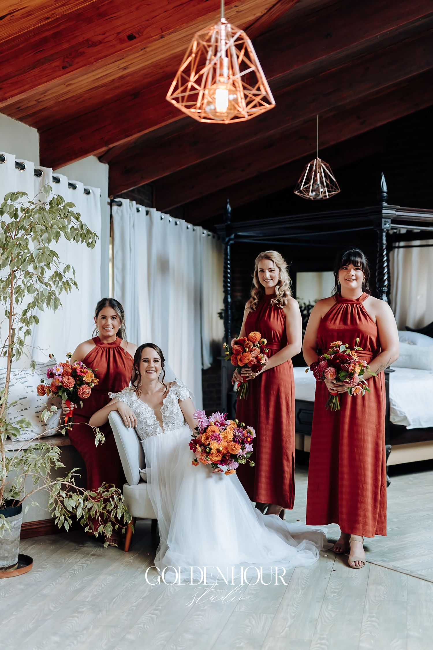 Bride in white lace gown seated with three bridesmaids in rust-red dresses holding vibrant bouquets indoors.