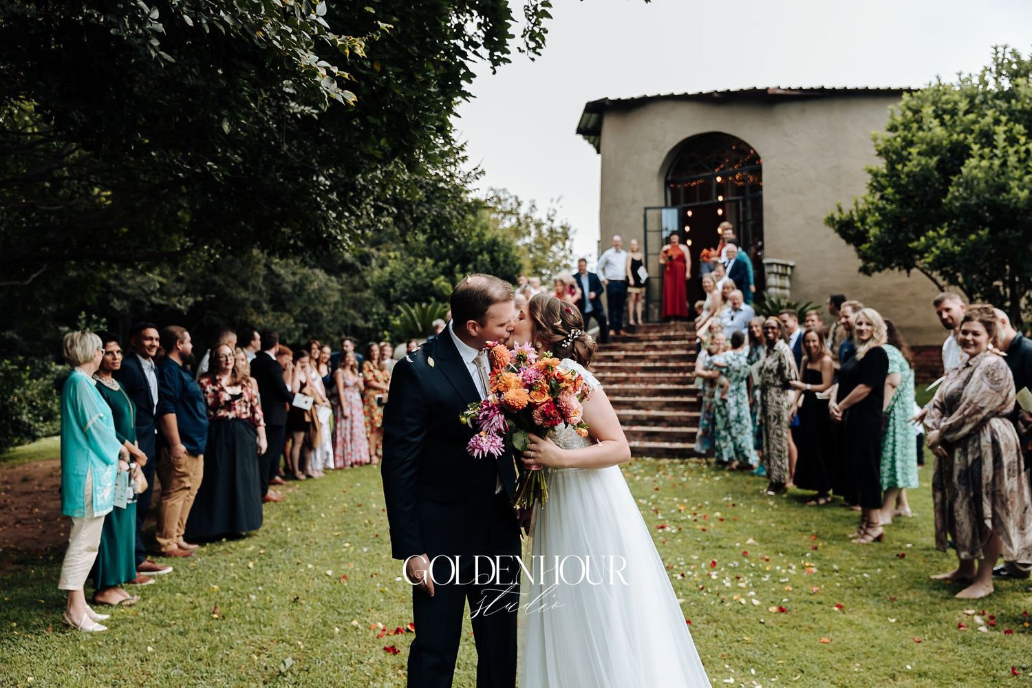 Bride and groom sharing first kiss as married couple before cheering wedding guests outdoors.