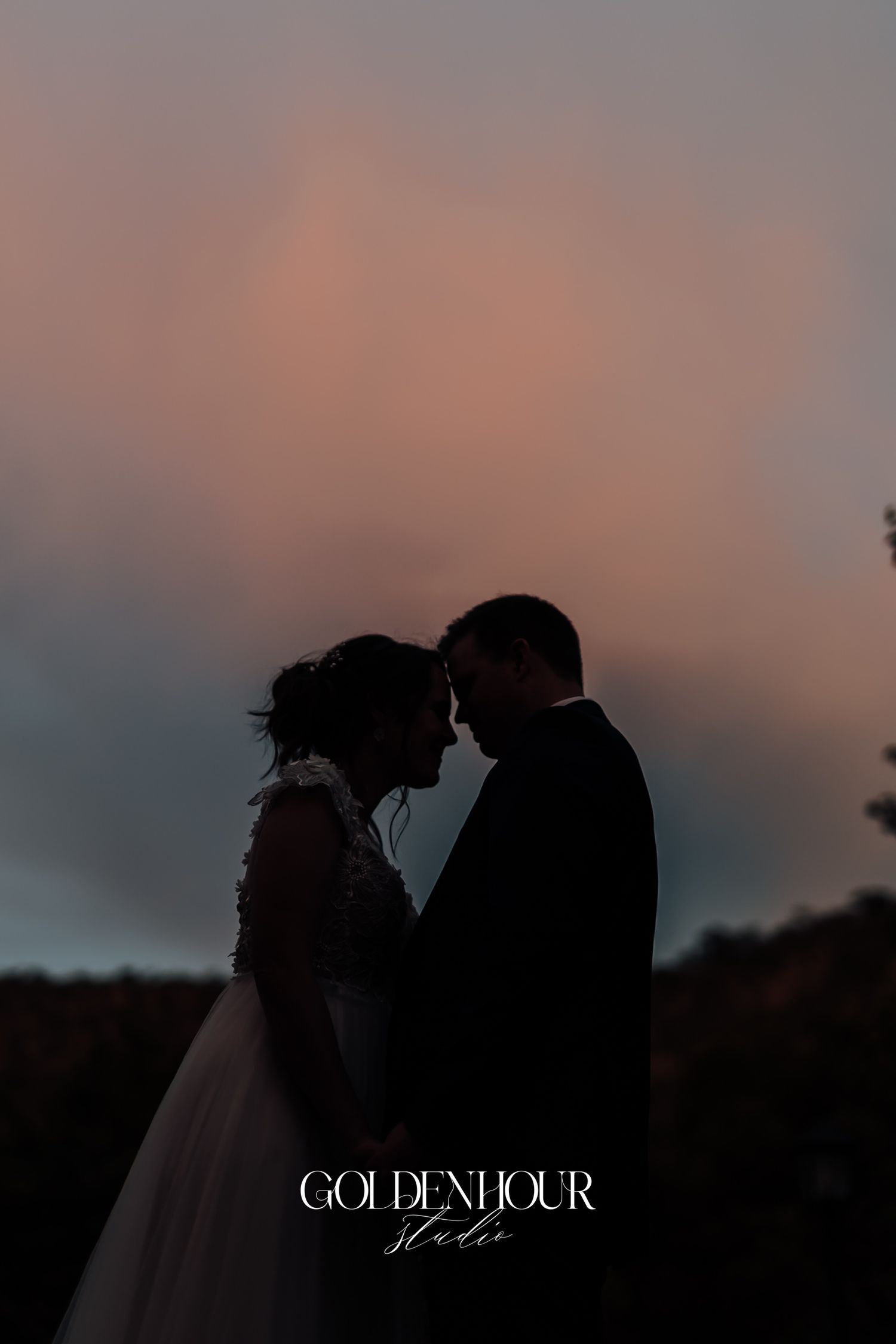 Romantic silhouette of bride and groom touching foreheads against a colorful pink and blue sunset sky.
