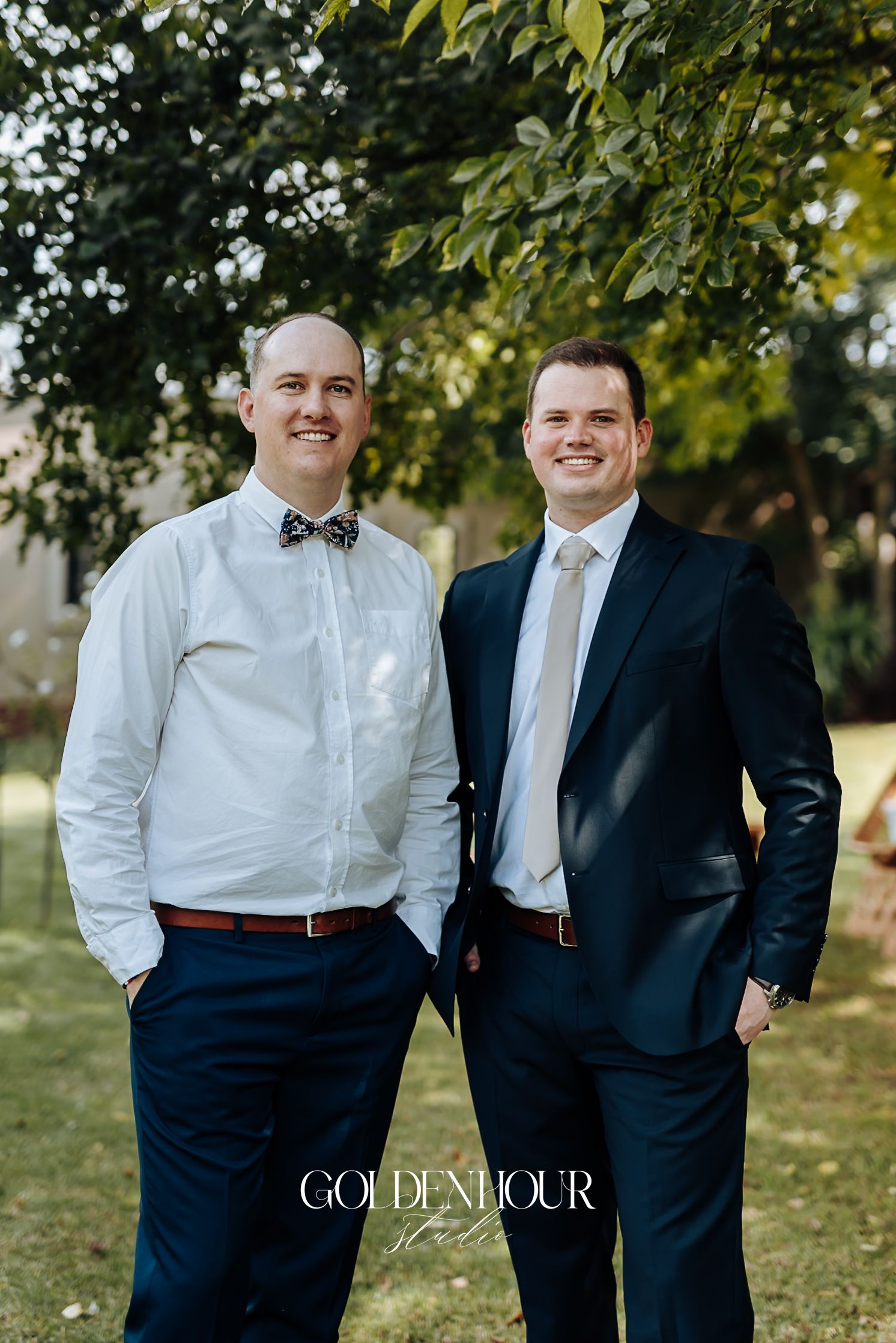 Two smiling groomsmen in navy suits posing outdoors under green trees at a wedding.