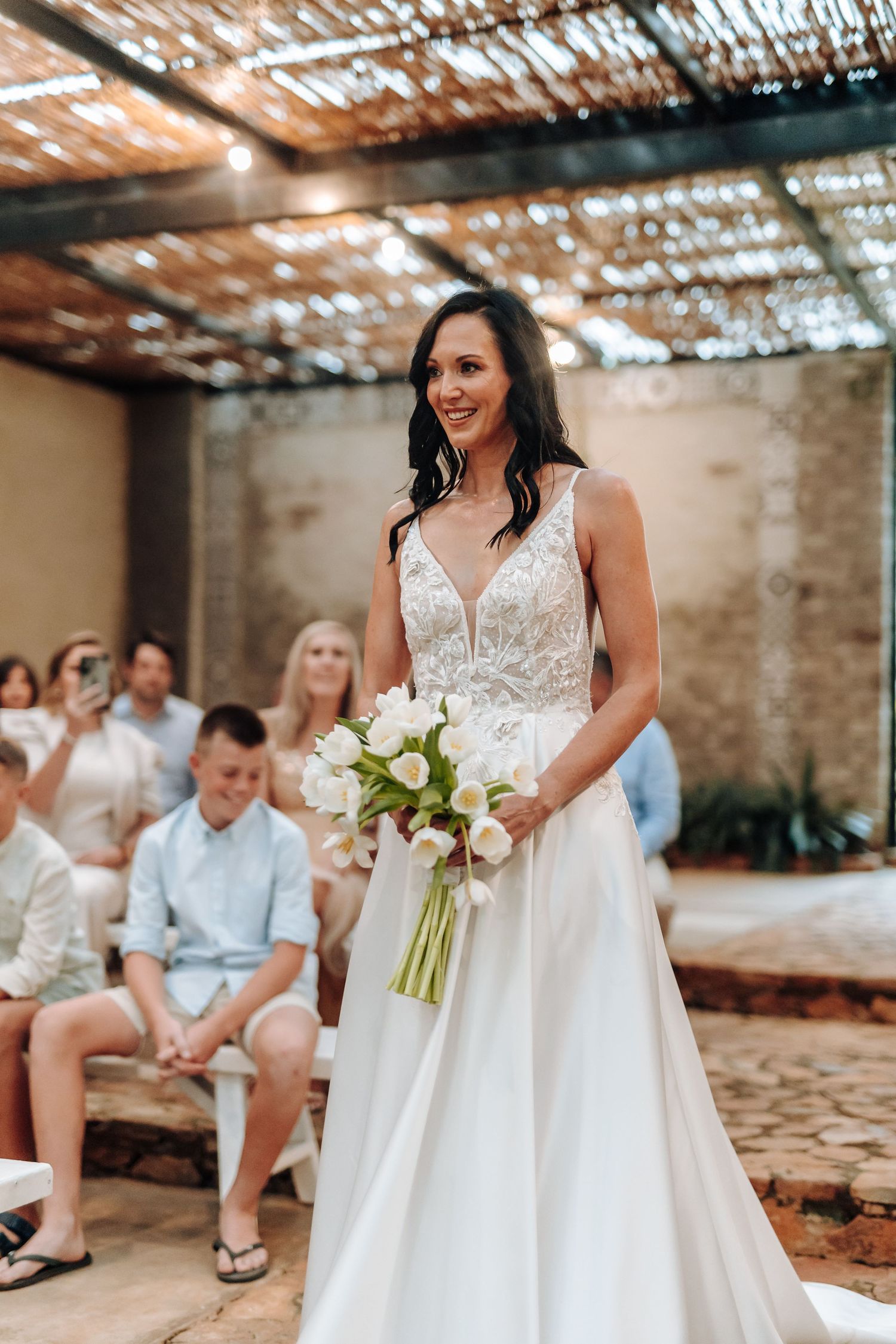 Smiling bride in a white lace gown holding a tulip bouquet walks down the aisle at an outdoor wedding.