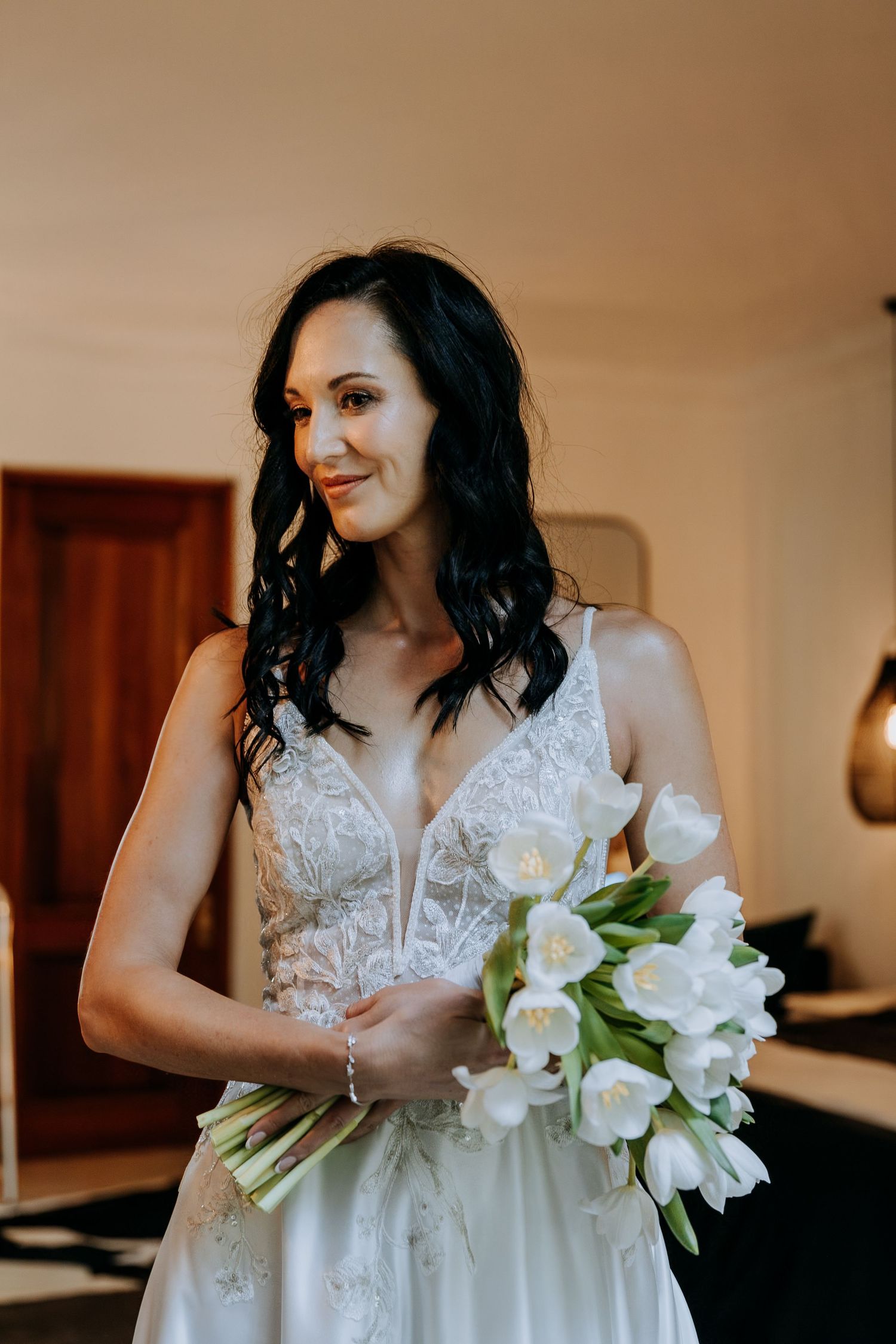 Bride in white lace gown holding elegant white tulip bouquet, smiling softly in warmly lit room.