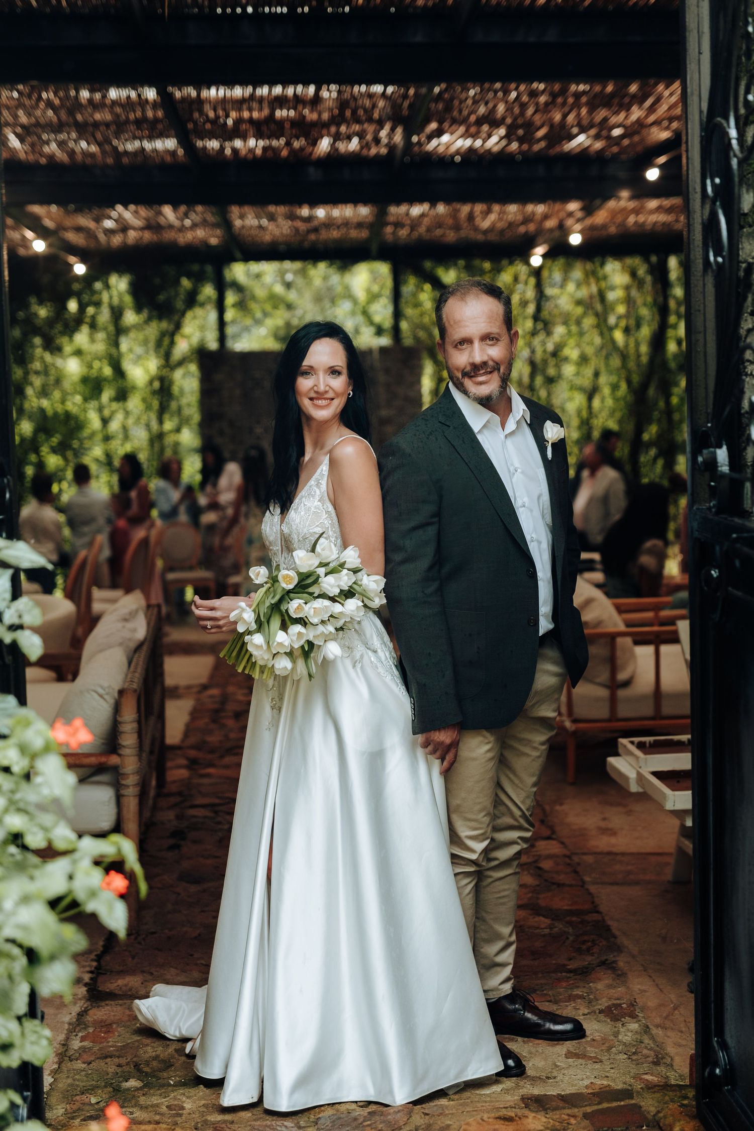 Bride in white gown holding tulip bouquet stands with groom in dark blazer at outdoor wedding venue.