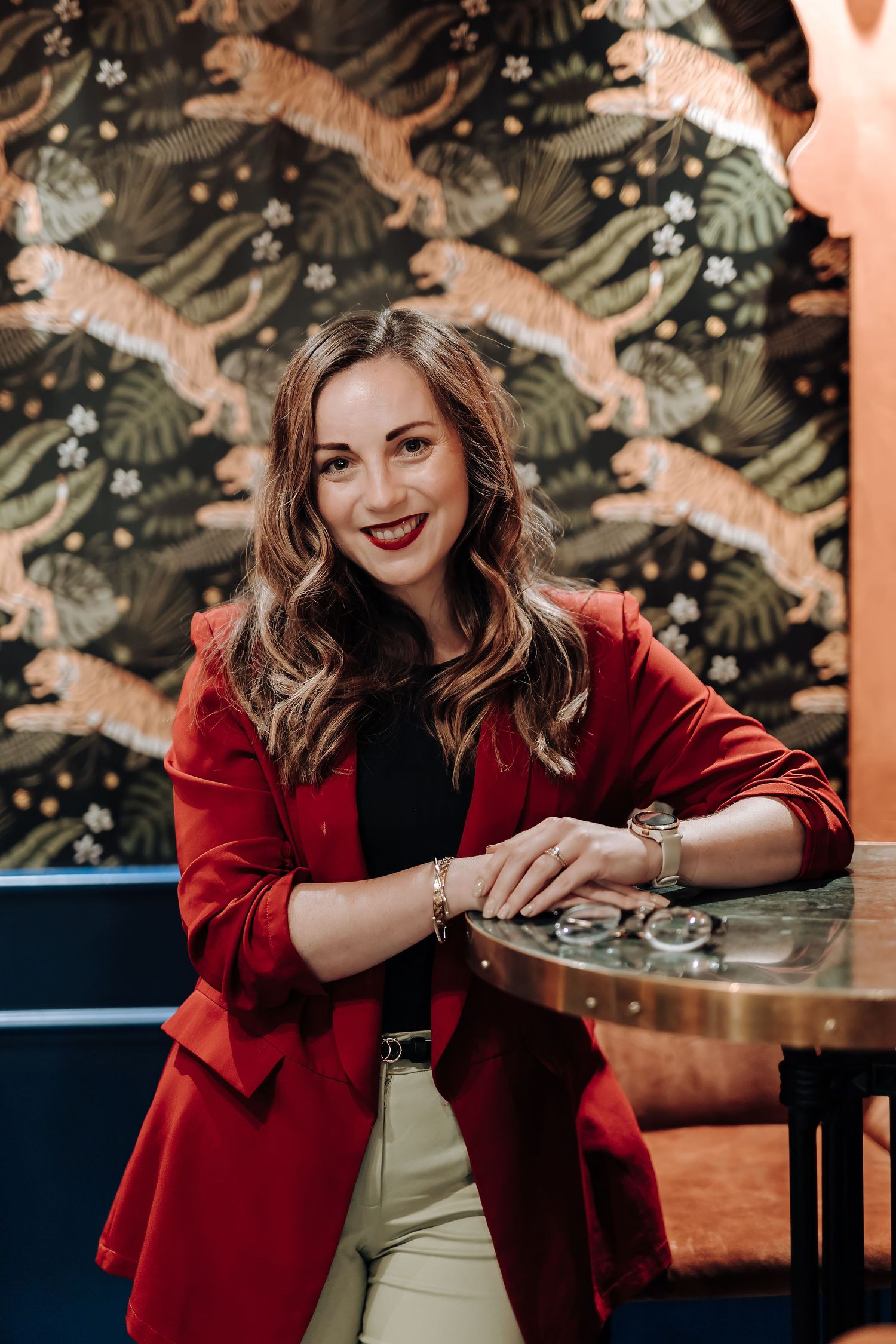Woman in vibrant red blazer sits at restaurant table with tropical foliage decor in background.