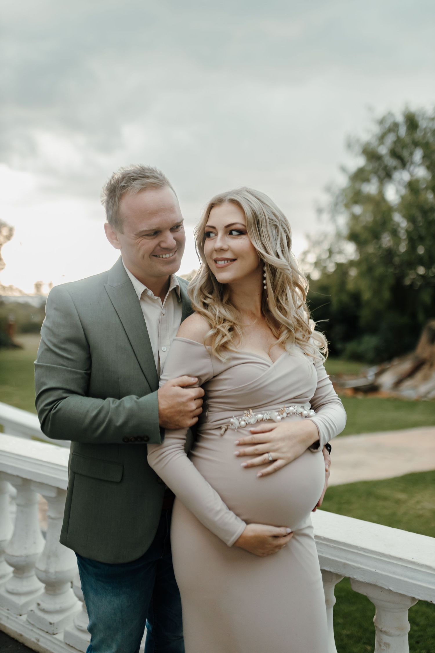 A couple shares an intimate moment on a white balustrade during an outdoor maternity photoshoot.
