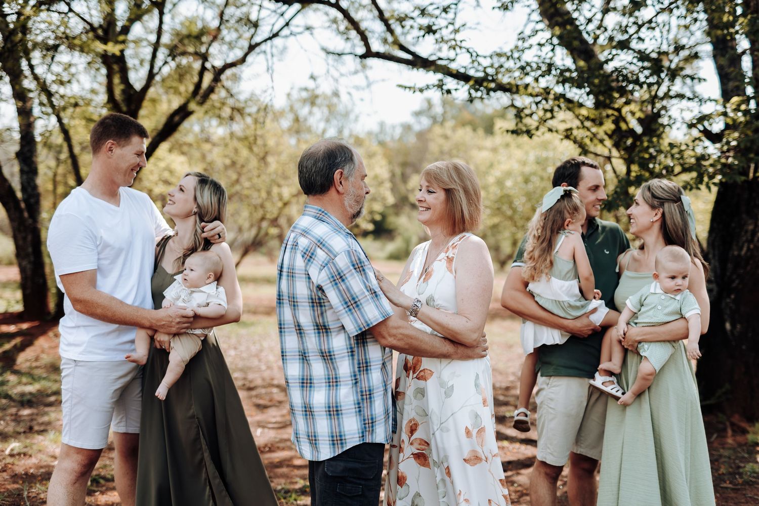 A family gathering outdoors under trees with multiple groups standing together in casual attire.