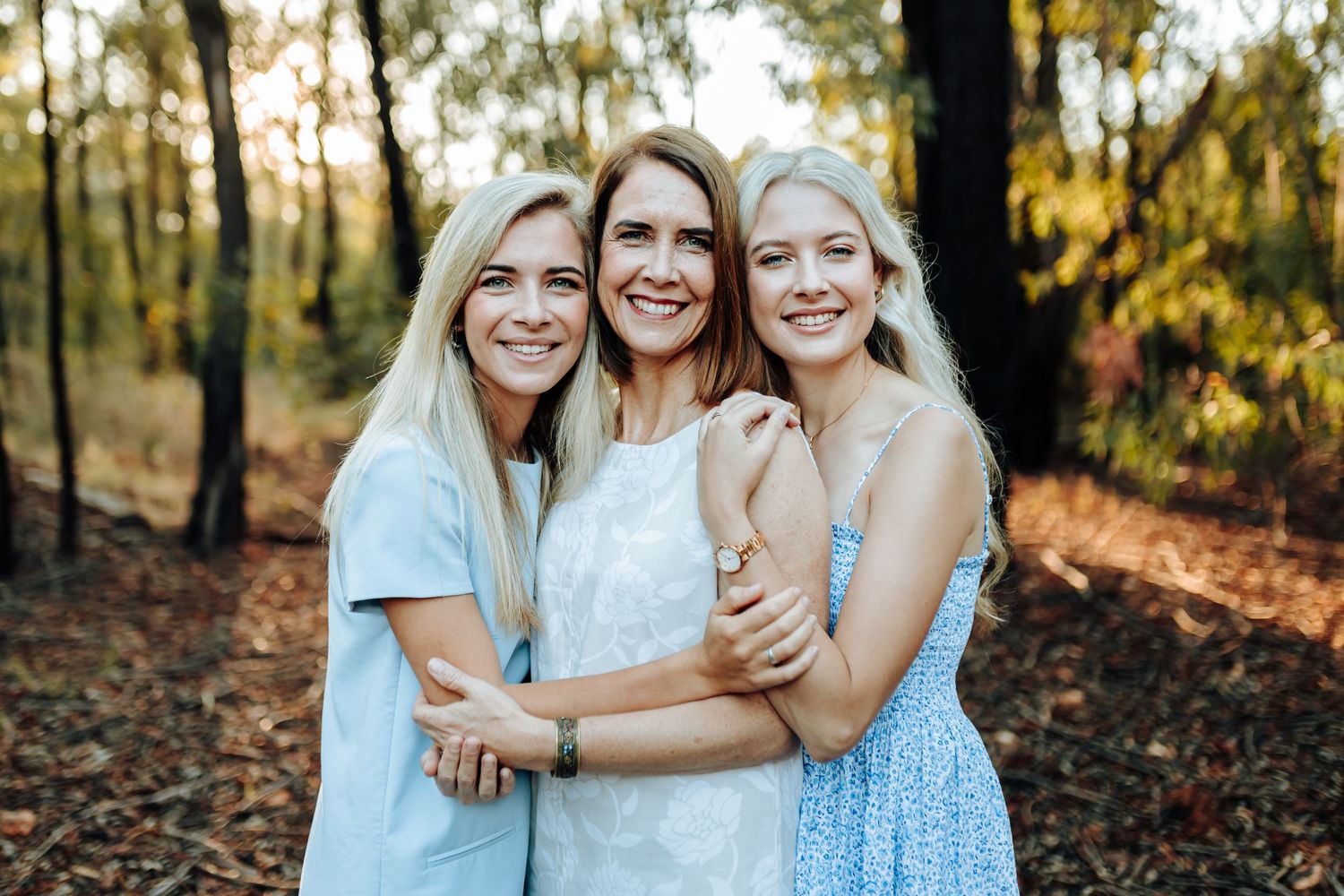 Family wearing coordinated blue and white outfits pose together during golden hour photoshoot in wooded setting.