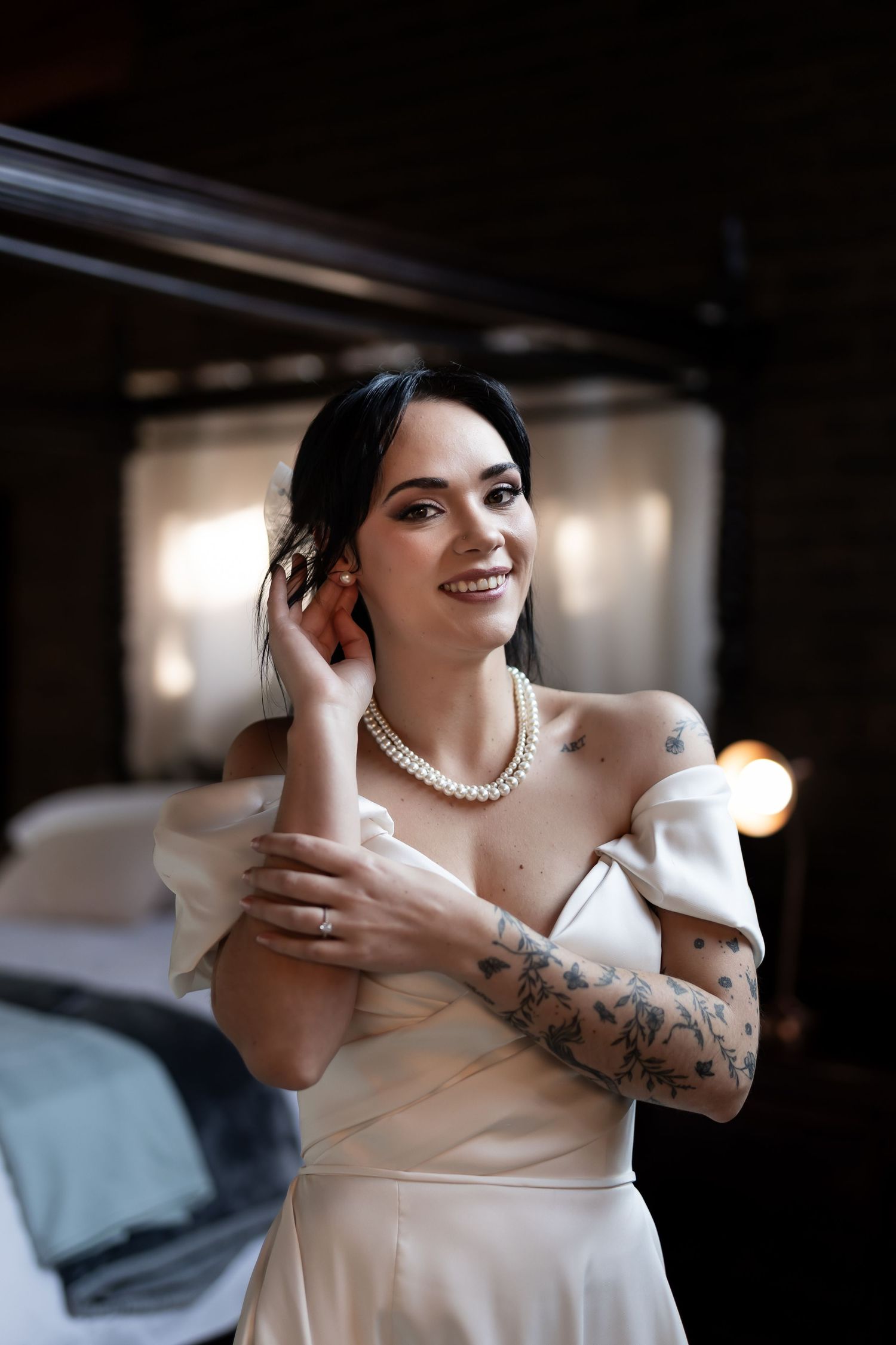 Elegant bride in off-shoulder cream dress with pearl necklace and arm tattoos smiles while getting ready in dim room.