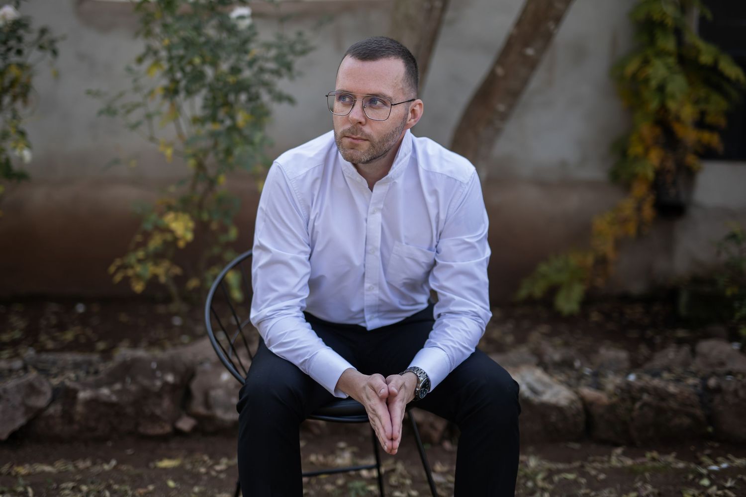 Person in glasses and white dress shirt sits thoughtfully on bench in garden setting with autumn foliage.