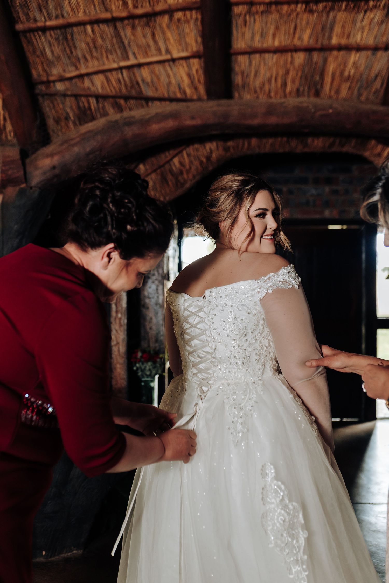 A wedding dress fitting scene under a rustic thatched roof with natural lighting and elegant lace details.
